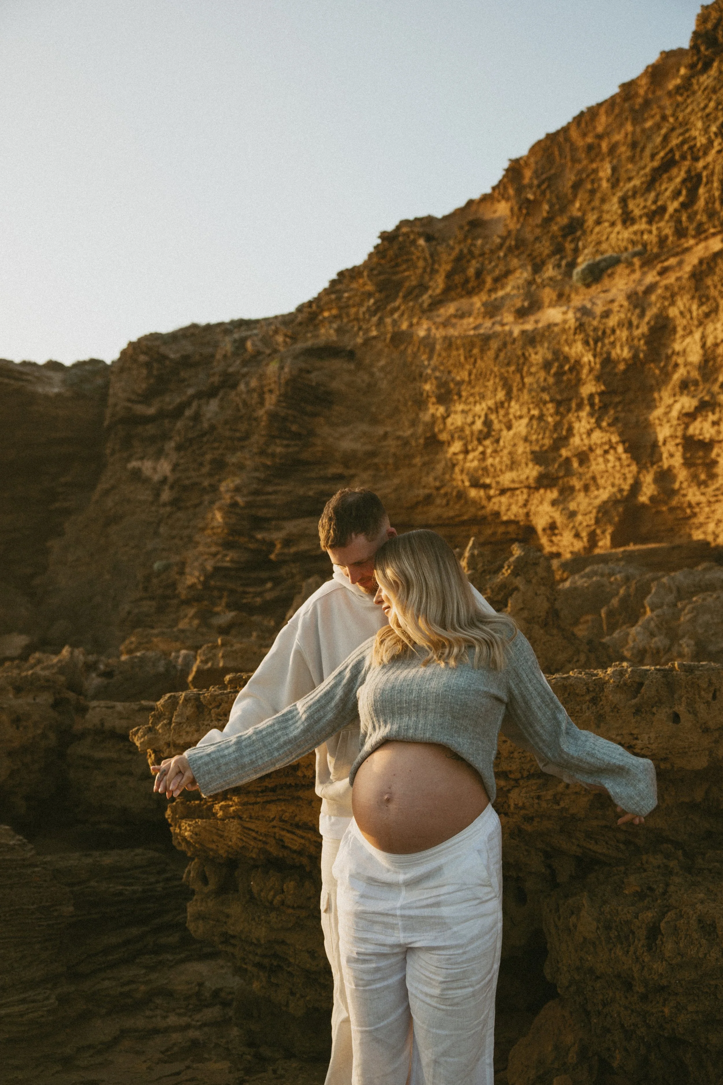 A pregnant woman and a man holding hands outdoors near rocky cliffs during sunset.