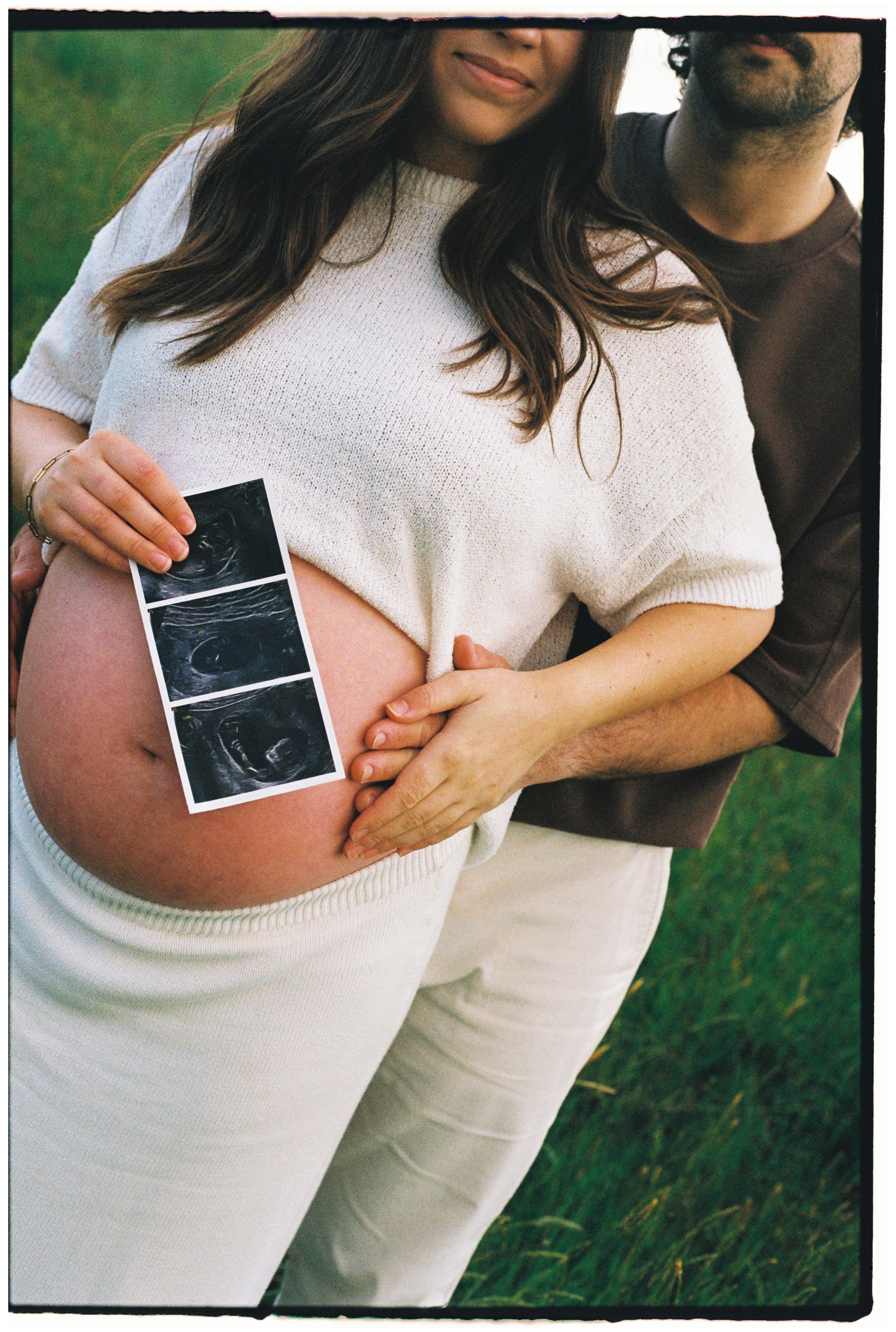 A pregnant woman dressed in a white sweater and pants, holding ultrasound images against her belly, standing outdoors on grass with an accompanying partner.