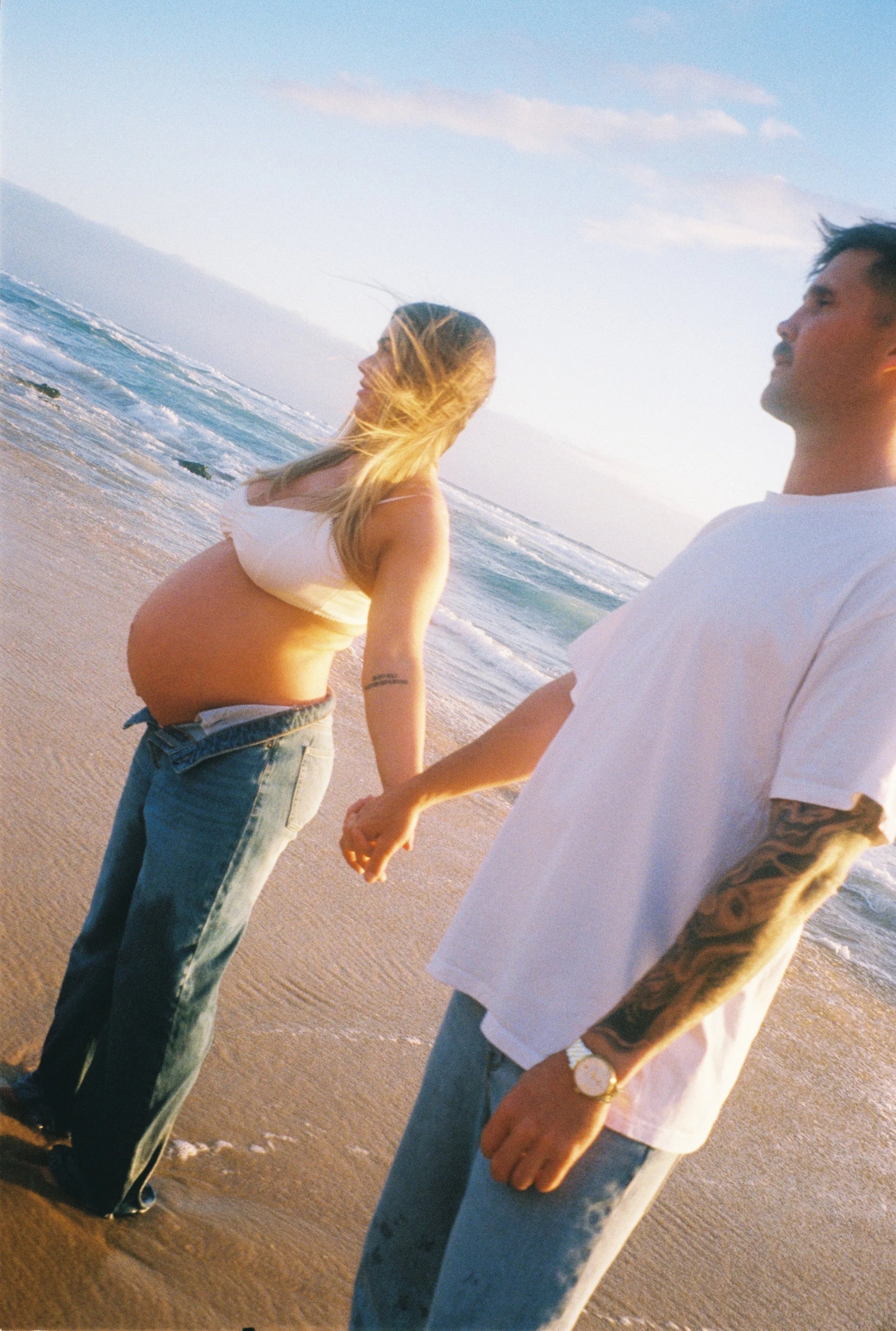 A pregnant woman and a man holding hands on the beach during sunset, with ocean waves in the background.