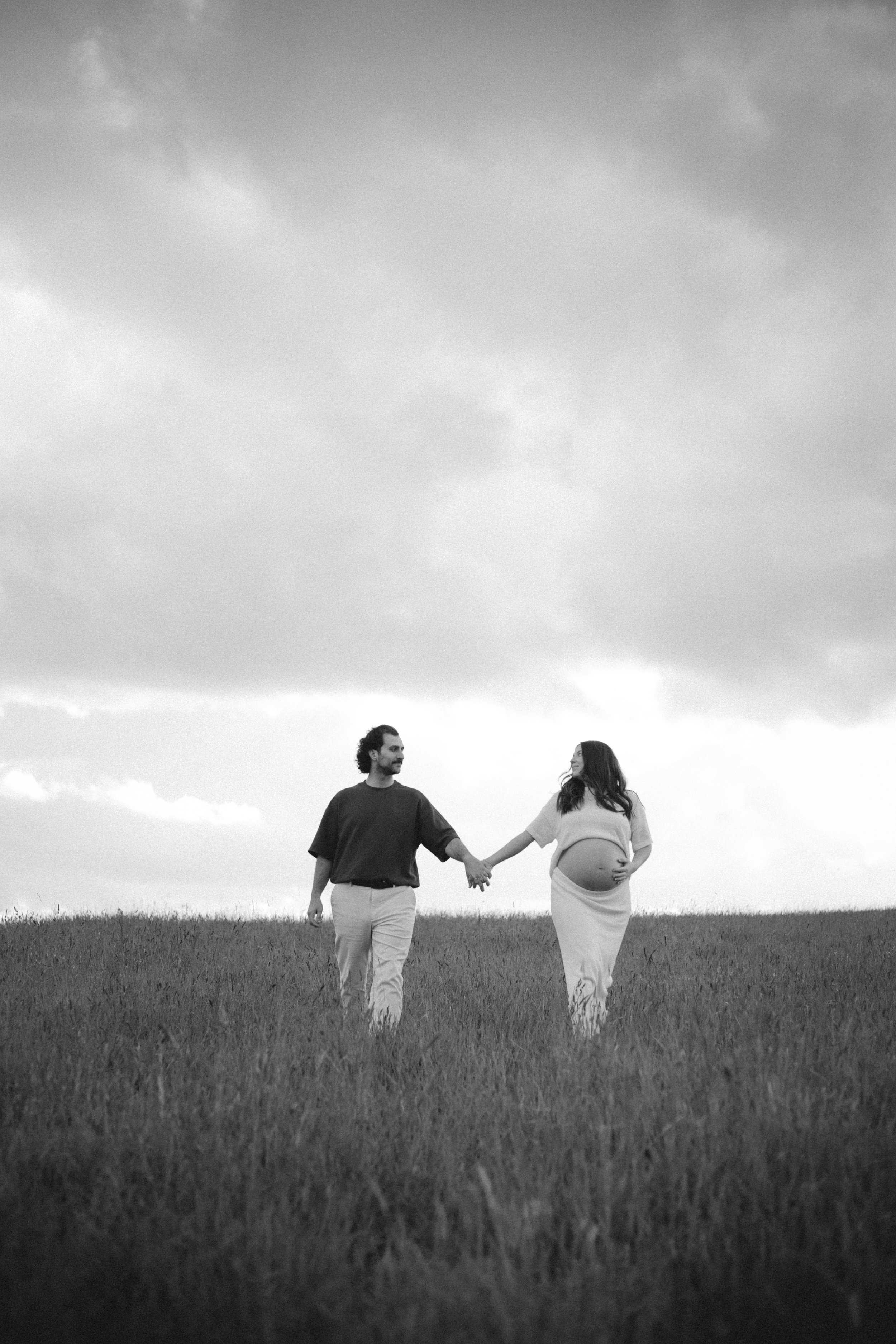 A pregnant woman and a man holding hands and walking in a grassy field under a cloudy sky.