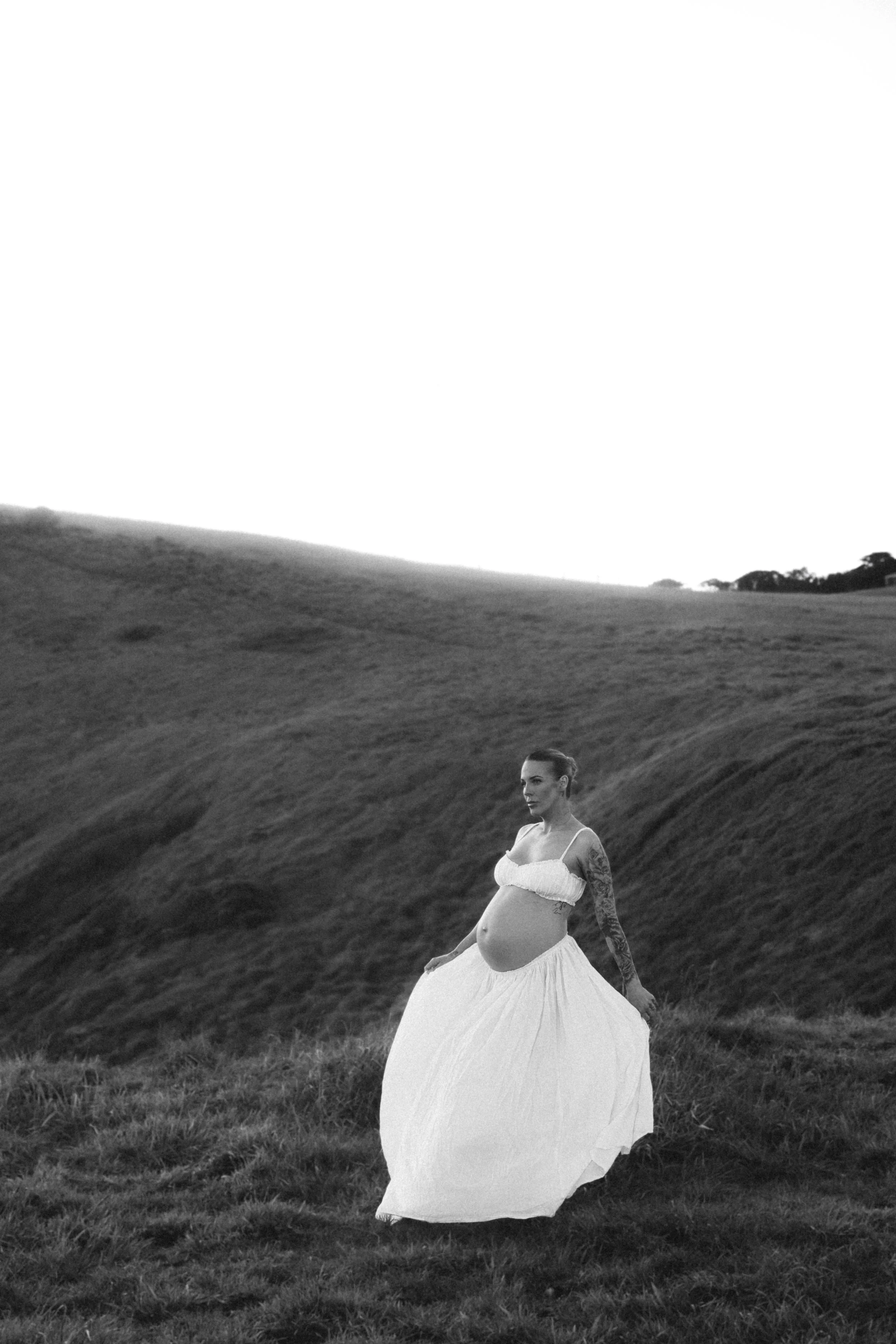 A pregnant woman in a white dress standing on a grassy hill in black and white.