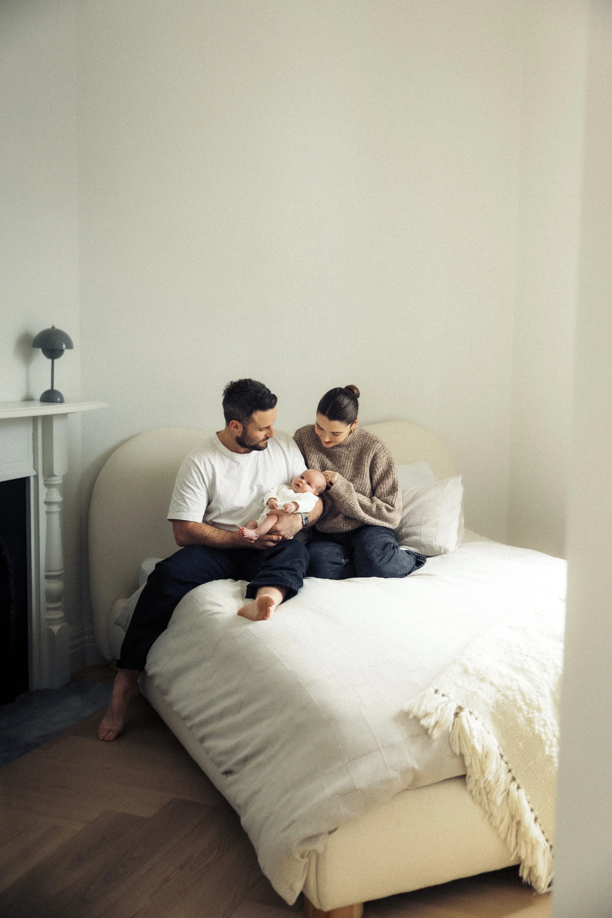 A couple sitting on a bed holding their baby, with the mother looking at the baby and the father holding the baby in his arms.