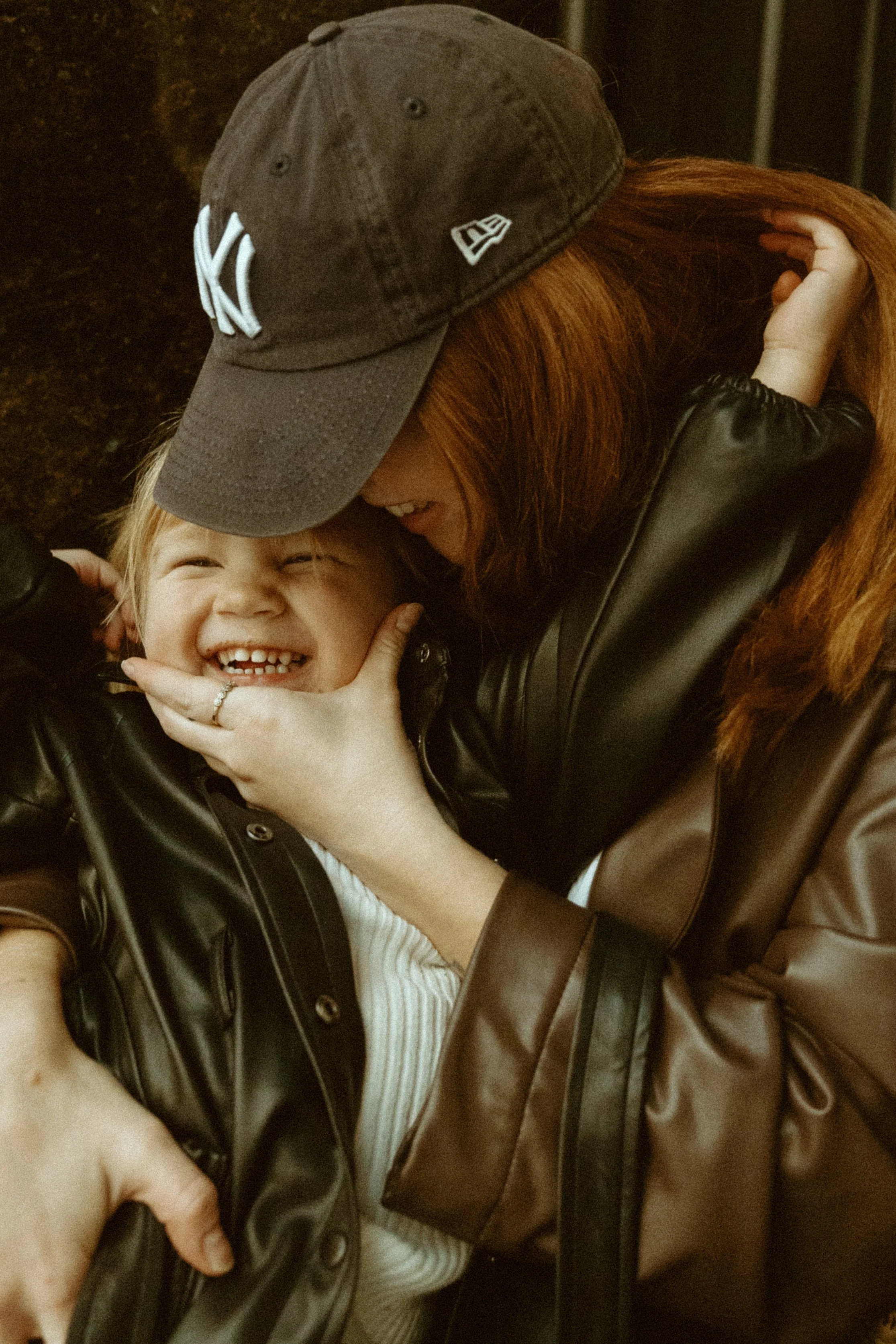 A woman with red hair wearing a New York Yankees baseball cap embraces and kisses a smiling young boy in a black leather jacket.