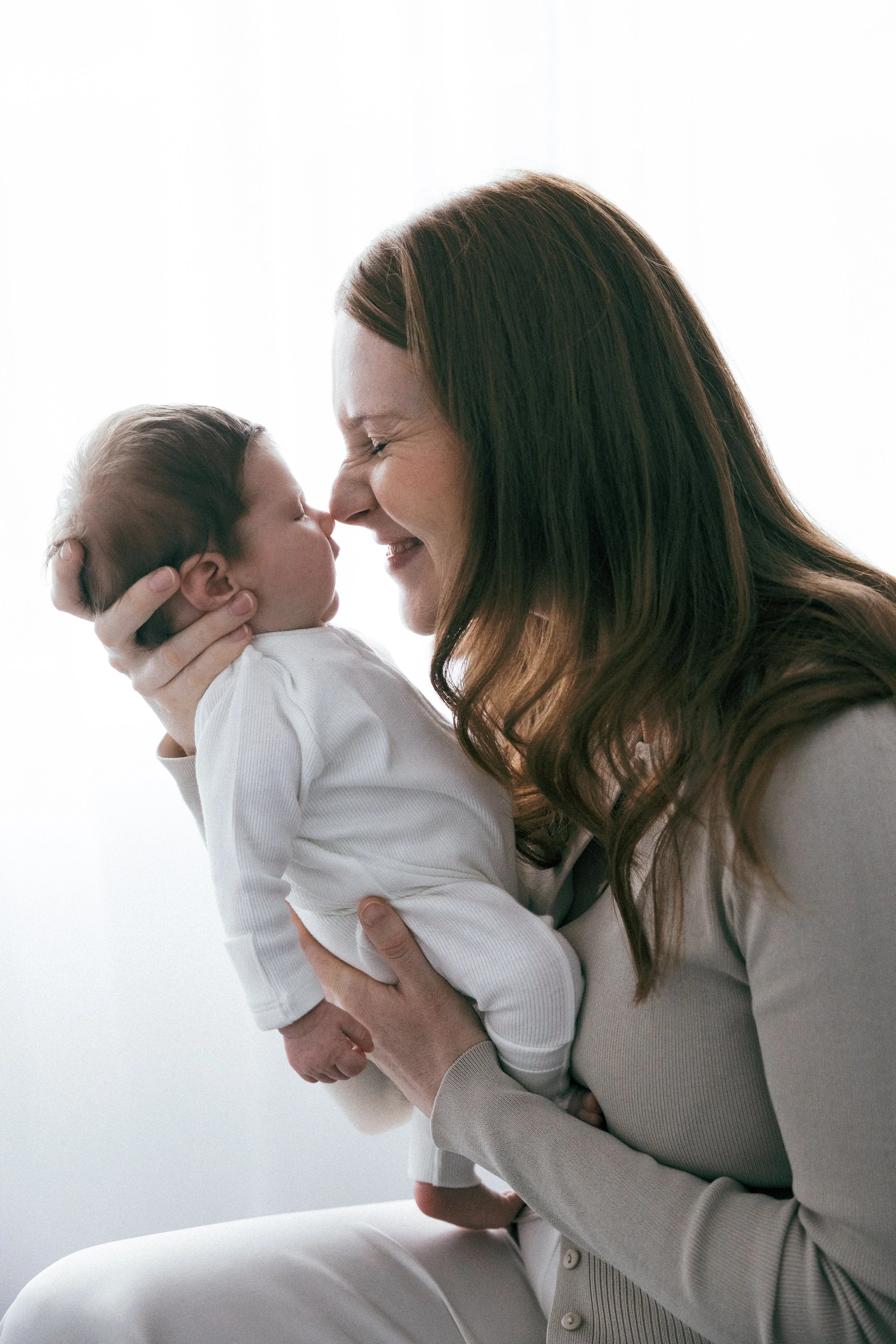 A woman holding a baby close, touching noses and smiling warmly, in front of a bright, white background.