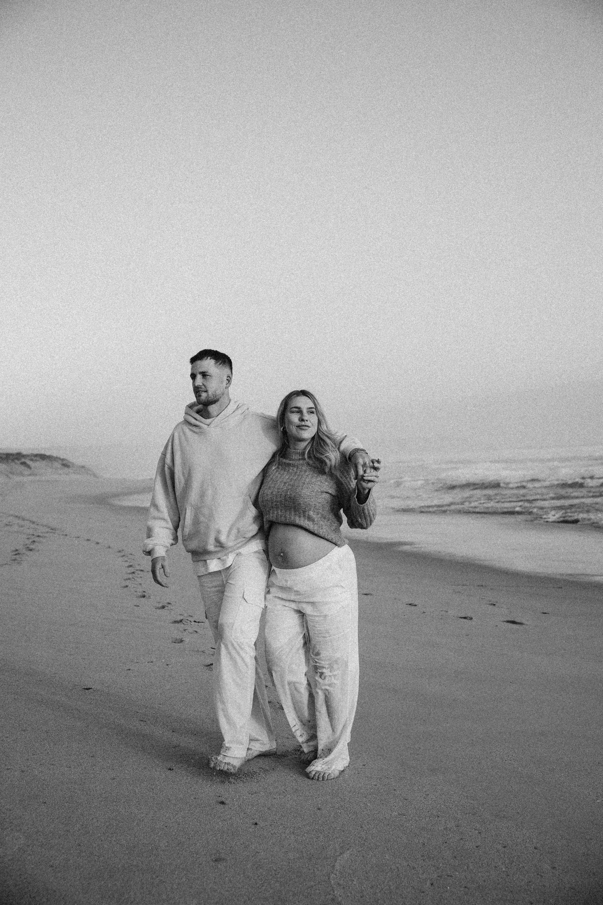 A couple walking on the beach, with the woman showing her pregnant belly, in black and white.