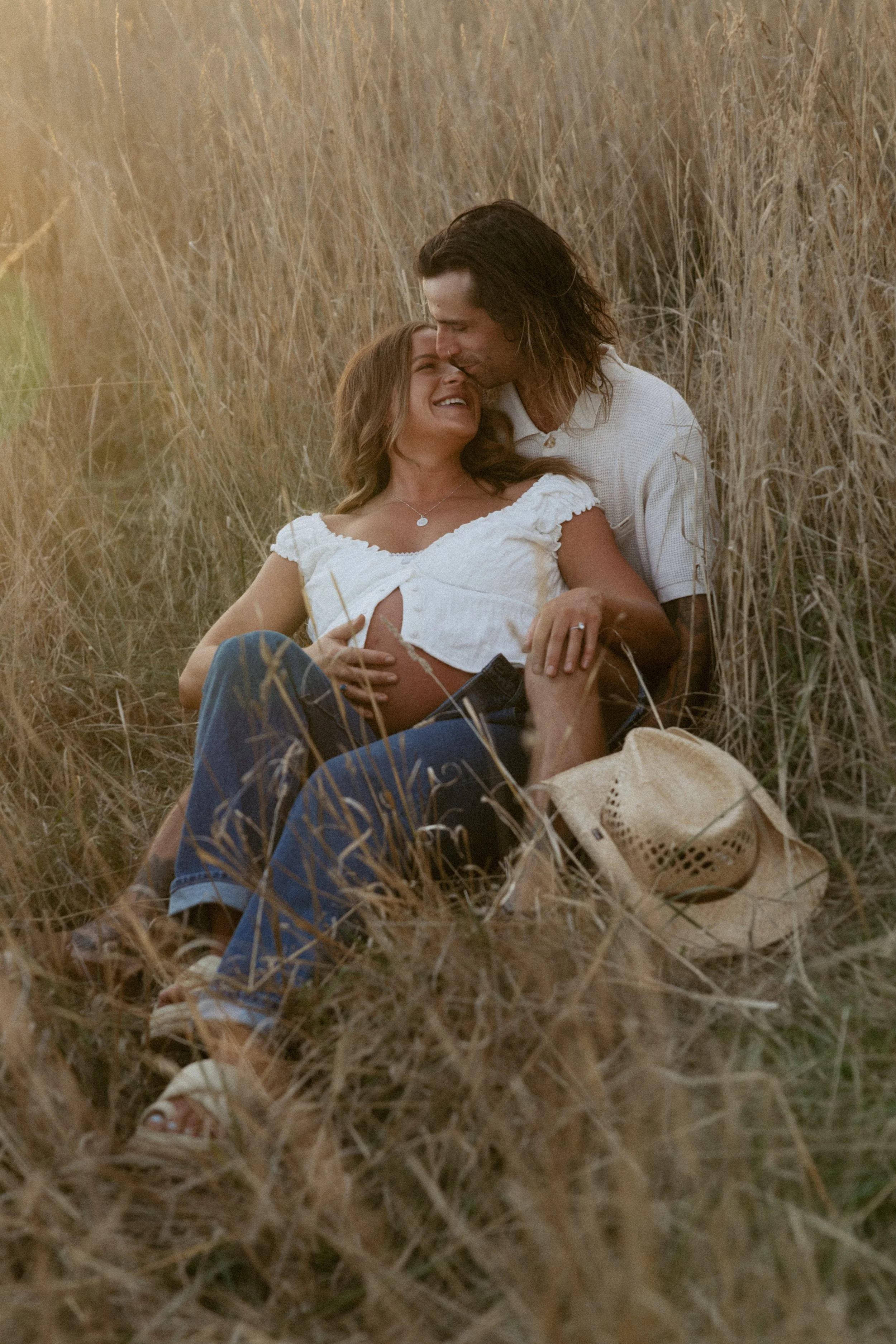 A couple sitting in a field of tall, golden grass during sunset, with the woman visibly pregnant, smiling and leaning into the man who is kissing her forehead.