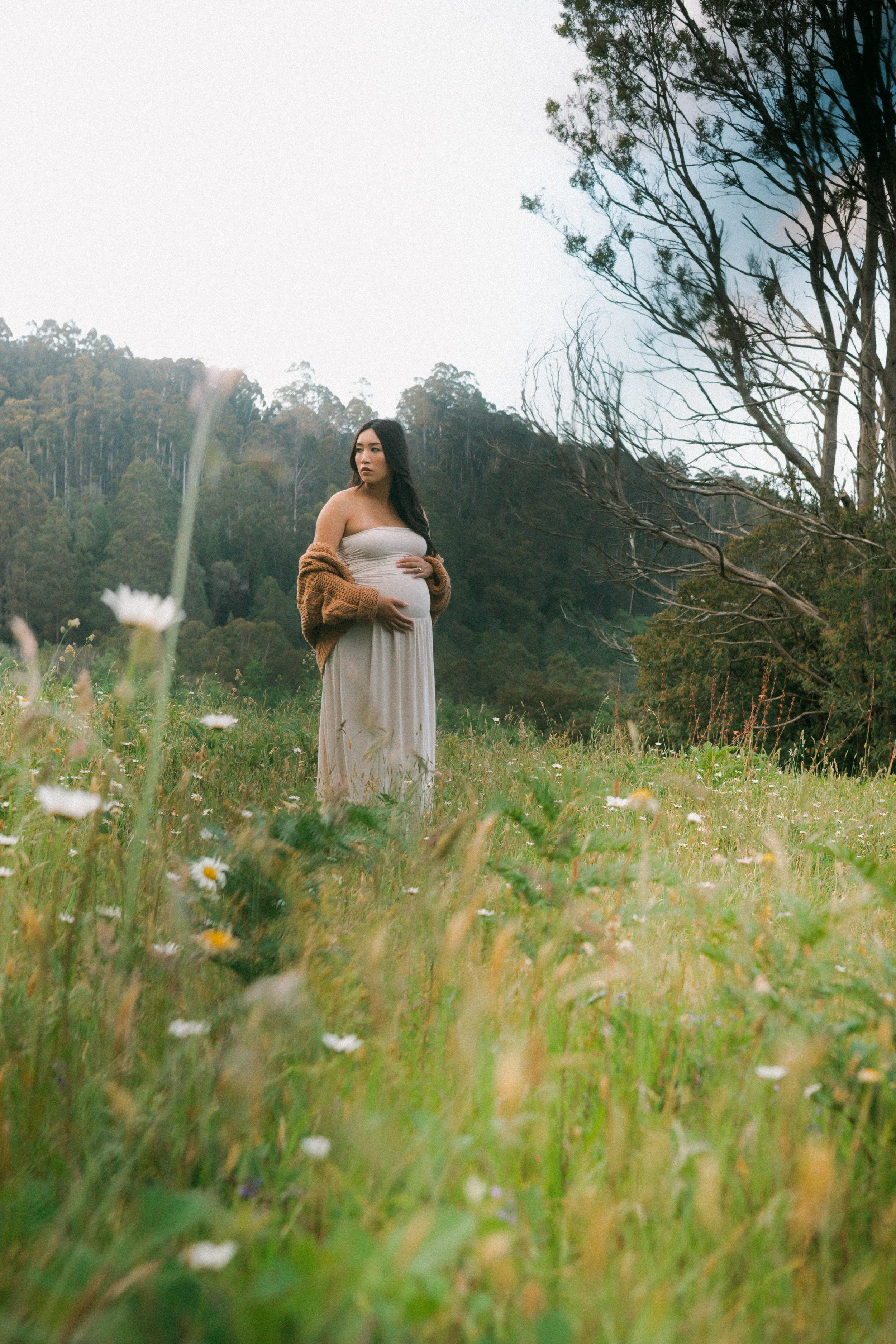 Pregnant woman standing in a grassy field with wildflowers, wearing a light dress and a brown sweater, surrounded by trees and mountains in the background.