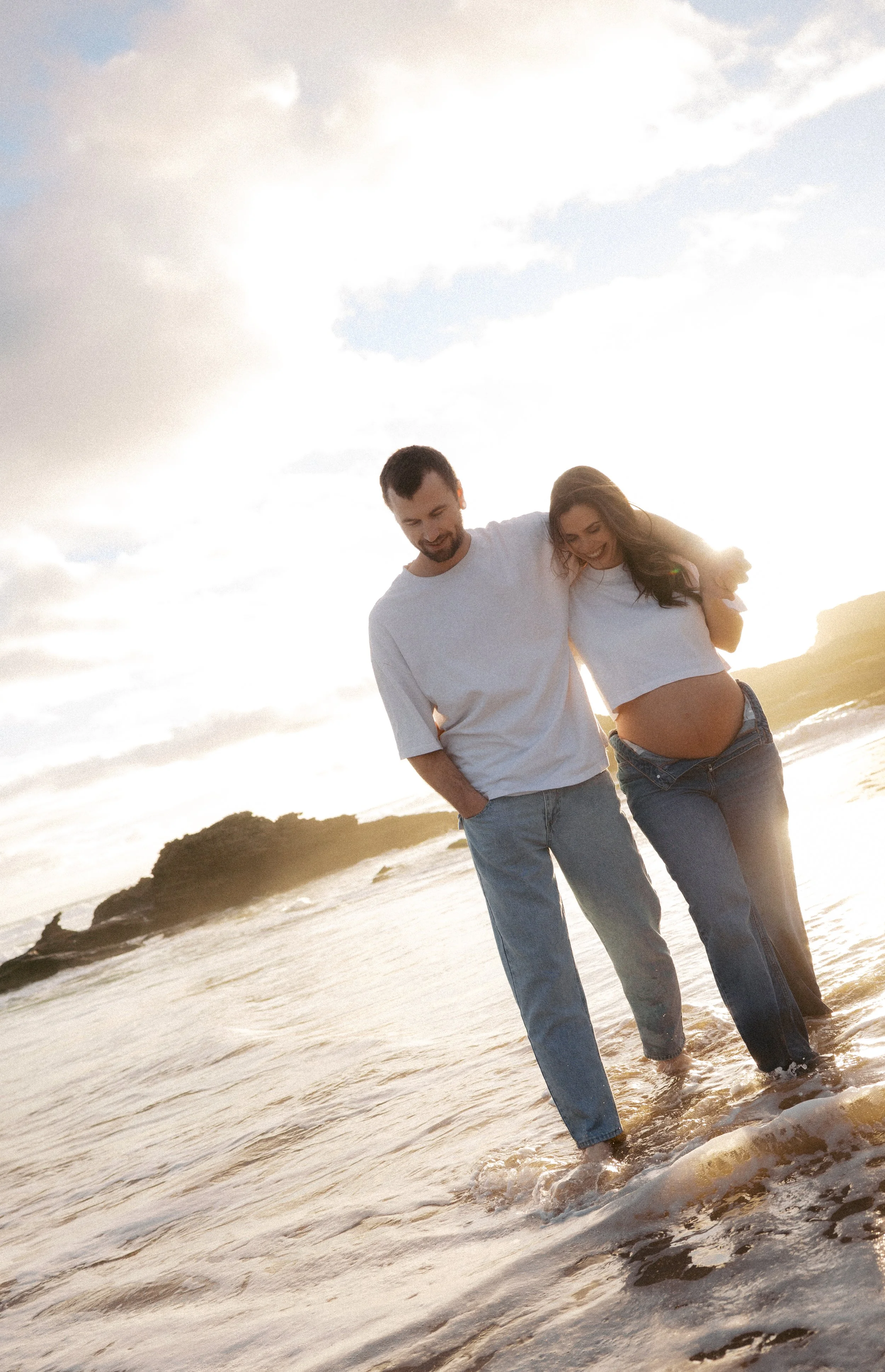 Pregnant woman and man walking in the ocean at sunset, smiling and enjoying the moment.
