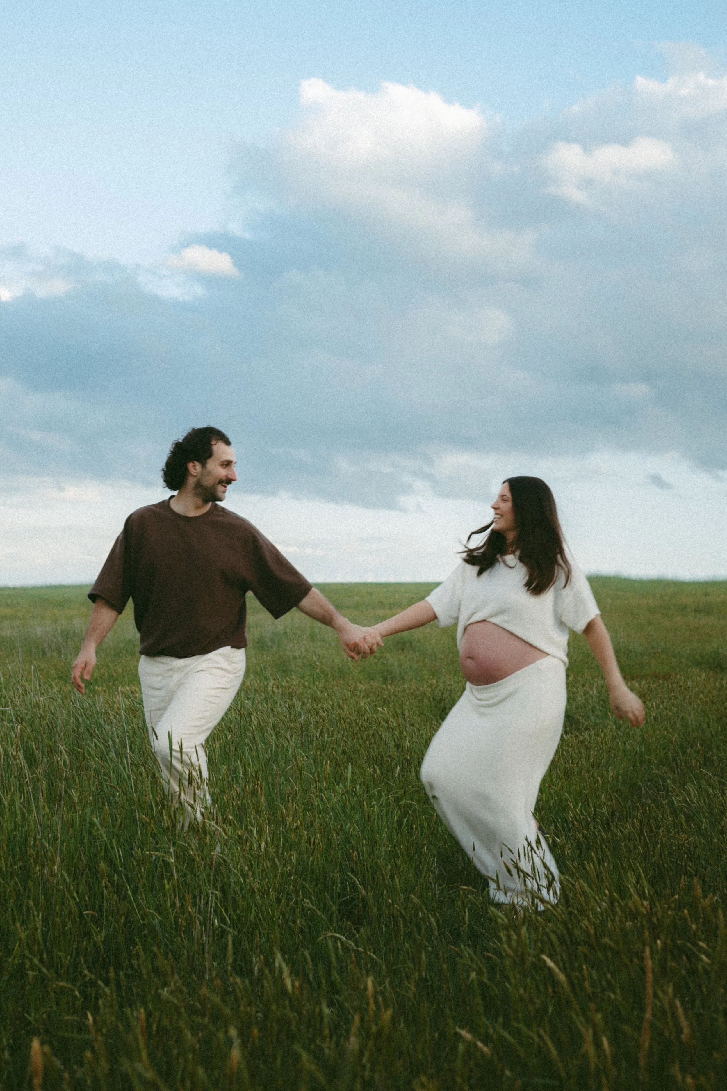 A pregnant woman and a man are holding hands and walking through a green field under a partly cloudy sky.
