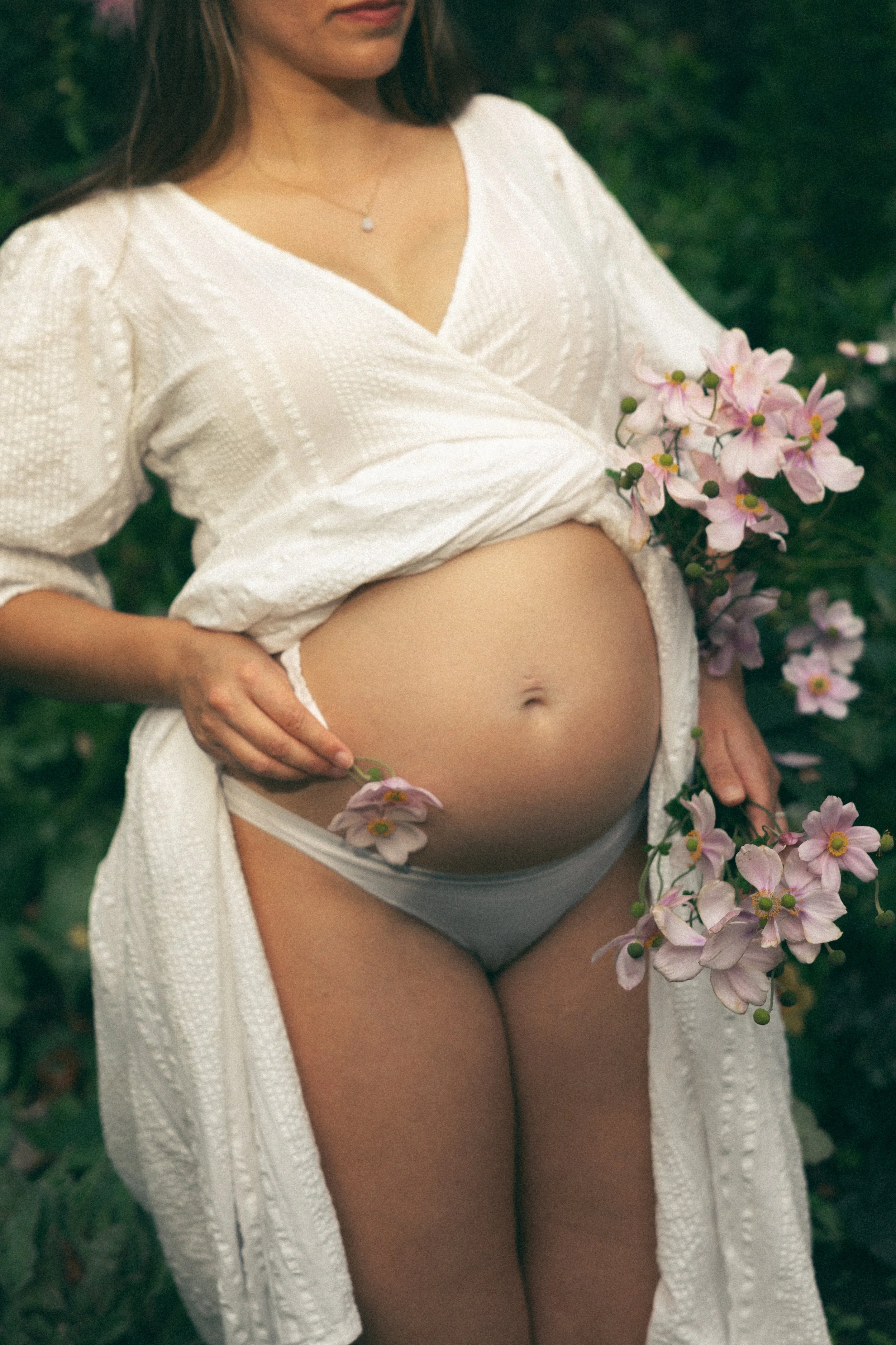 A pregnant woman standing outdoors holds pink flowers, wearing a white robe and white underwear.