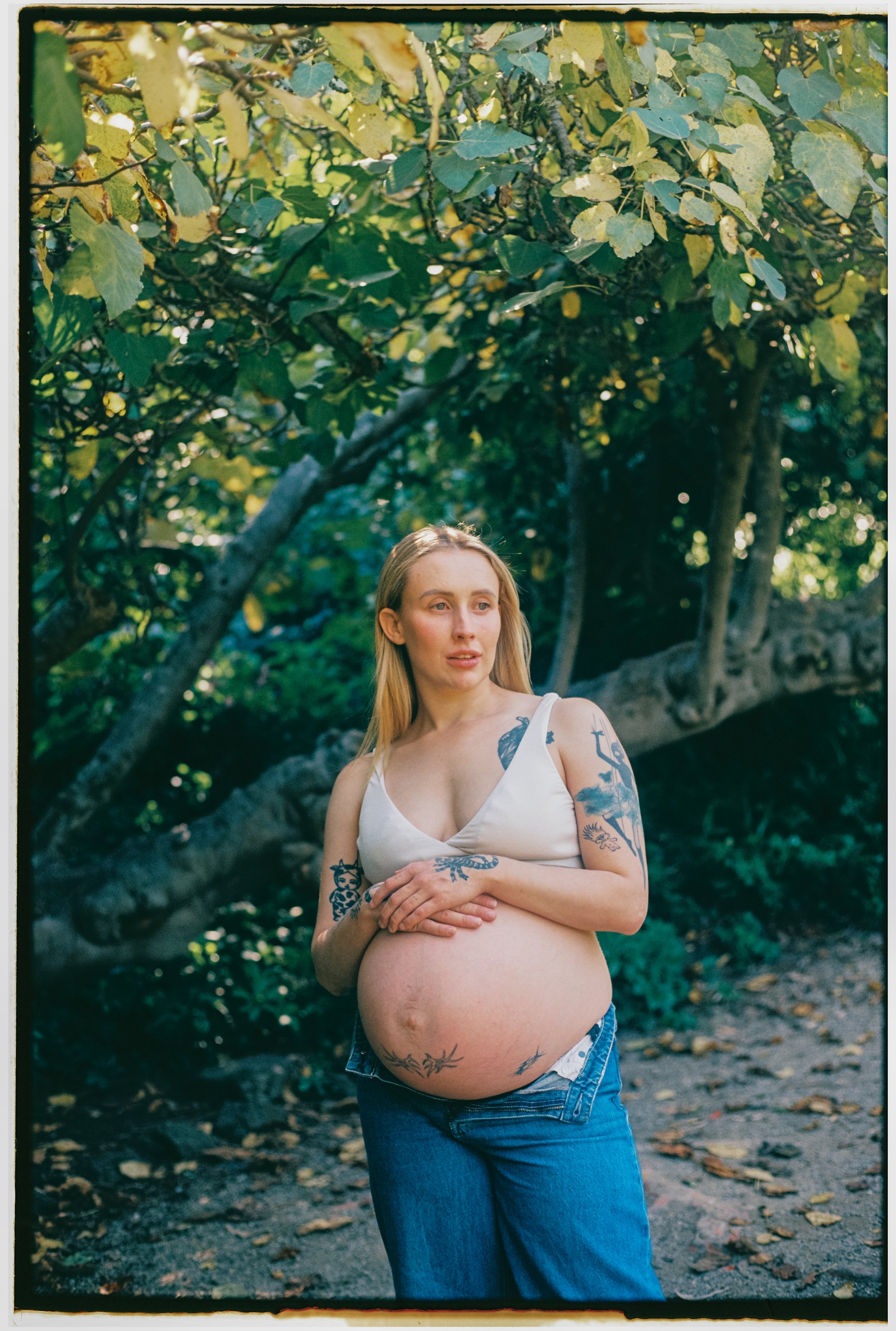 A pregnant woman with tattoos on her arms and a tattoo on her belly is standing outdoors, surrounded by trees and greenery, wearing a white tank top and blue jeans. Maternity, Newborn, Family & Couples Photography | Doreen, Melbourne
