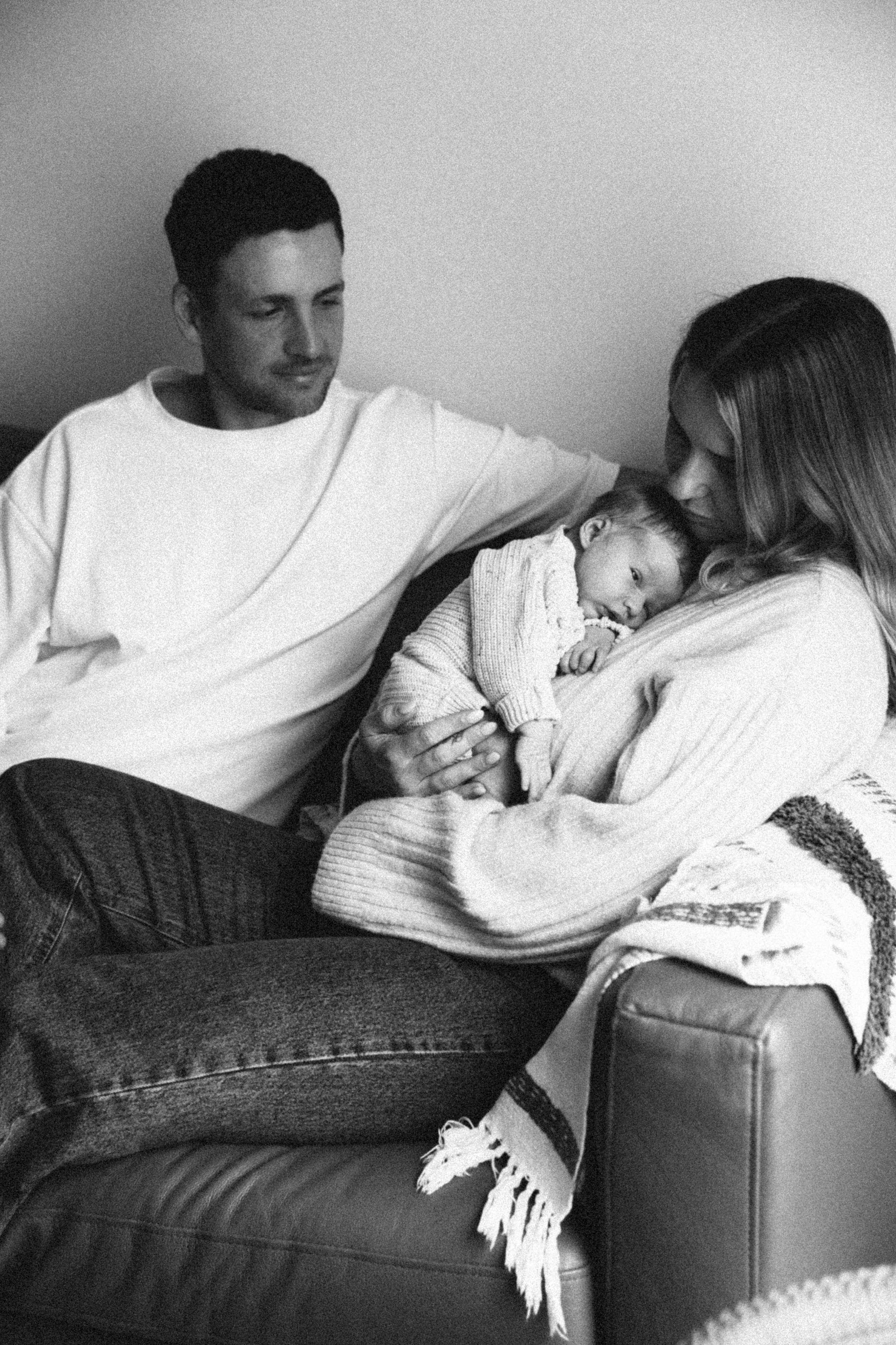 A black-and-white photo of a family with a mother, father, and their baby. The mother is holding the baby close to her chest while sitting on a couch. The father is sitting beside her with his arm around her shoulders.