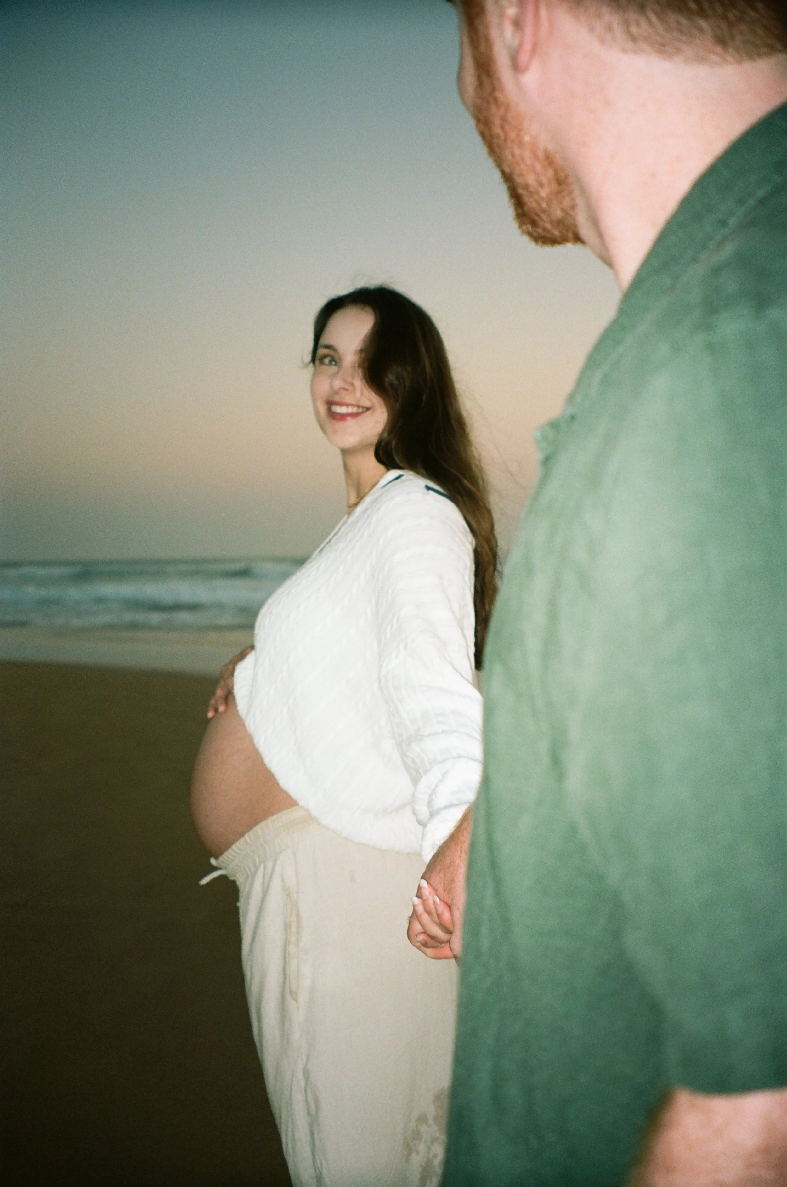 A pregnant woman smiling at the camera while holding hands with a man on a beach at sunset.