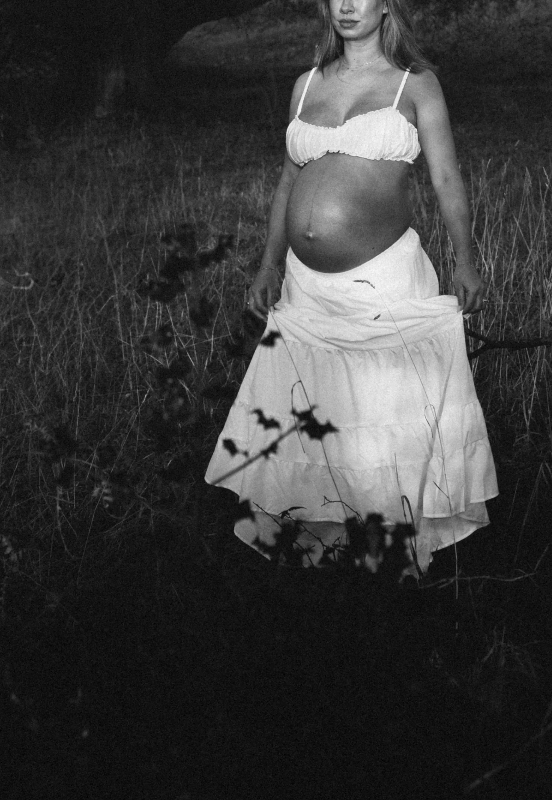 A pregnant woman in a white skirt and crop top standing in a grassy field, holding out her skirt, with some foliage in the foreground.