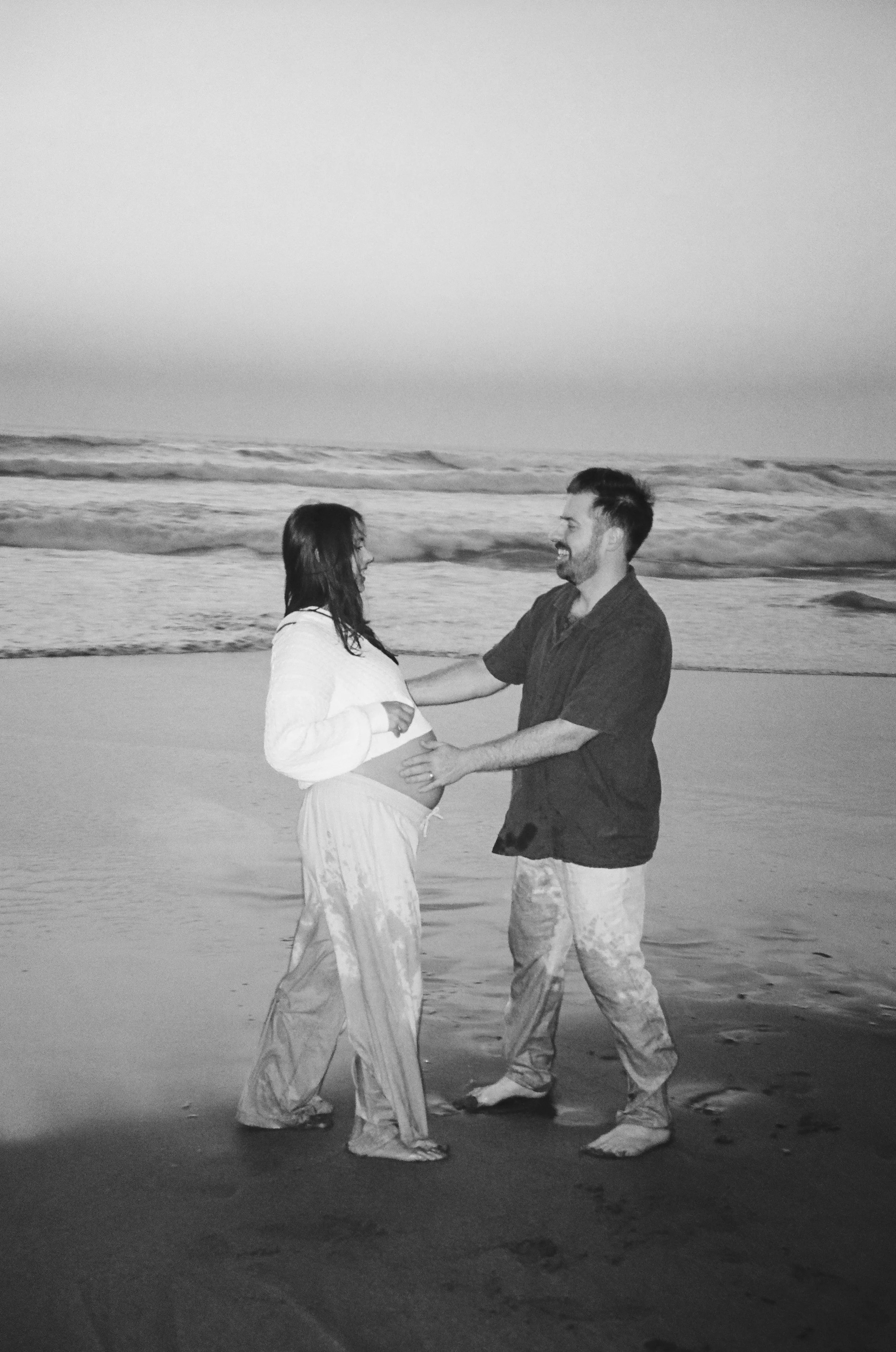 A black and white photo of a couple on the beach, with the woman visibly pregnant. They are facing each other, smiling, with the man holding her belly, and the ocean waves in the background.