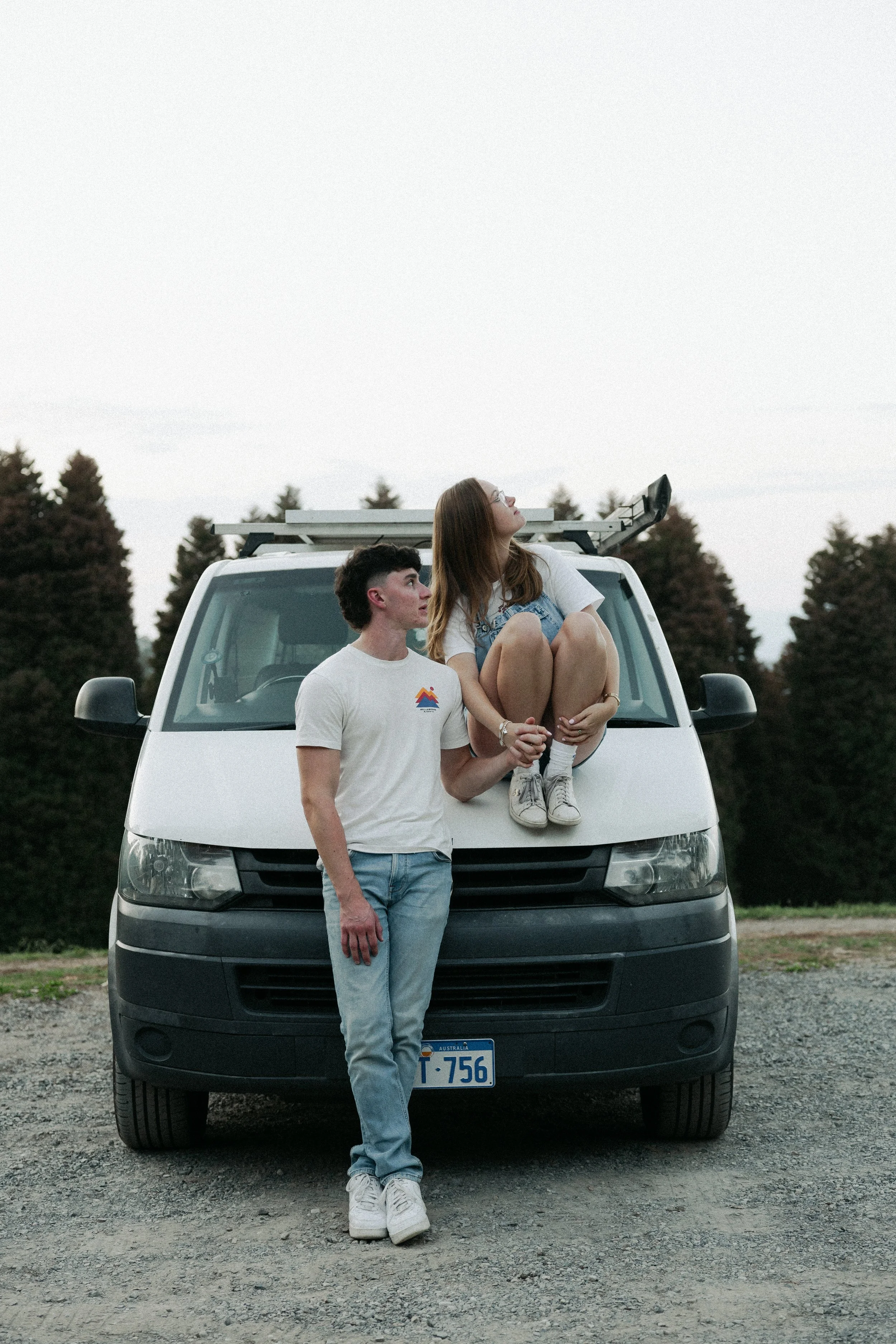 A young man and woman with light skin and brown hair standing and sitting on a white van with an Australian license plate. The woman is seated on the van's hood, holding hands with the man, who is standing close by. They are outdoors with trees in th