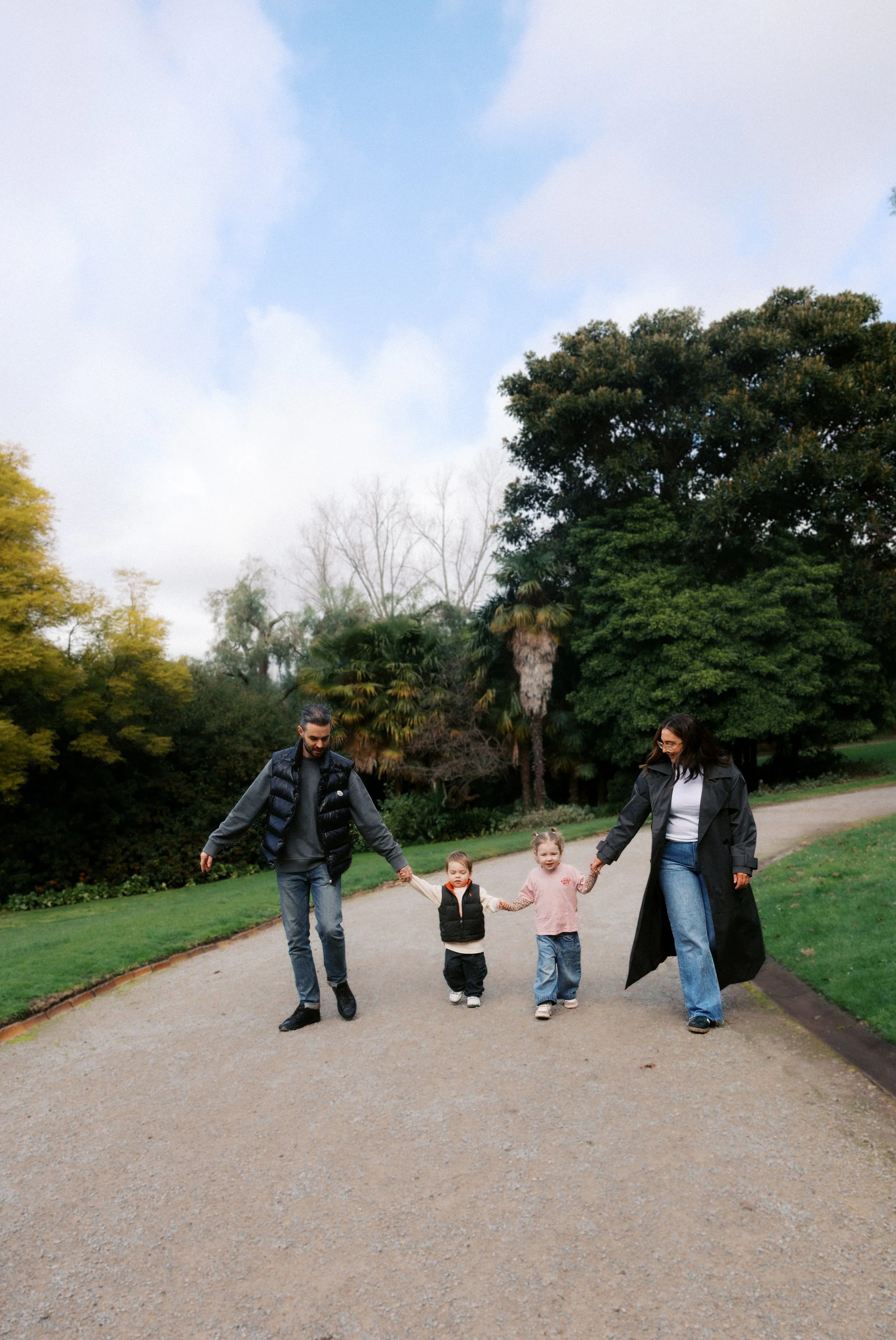 A family of four walking hand-in-hand down a park pathway with trees and cloudy sky in the background.