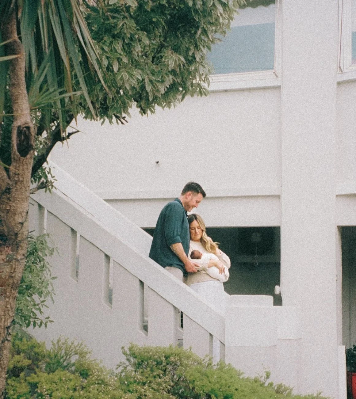 A couple stands on an outdoor staircase holding a newborn baby, with a white building and plants nearby.