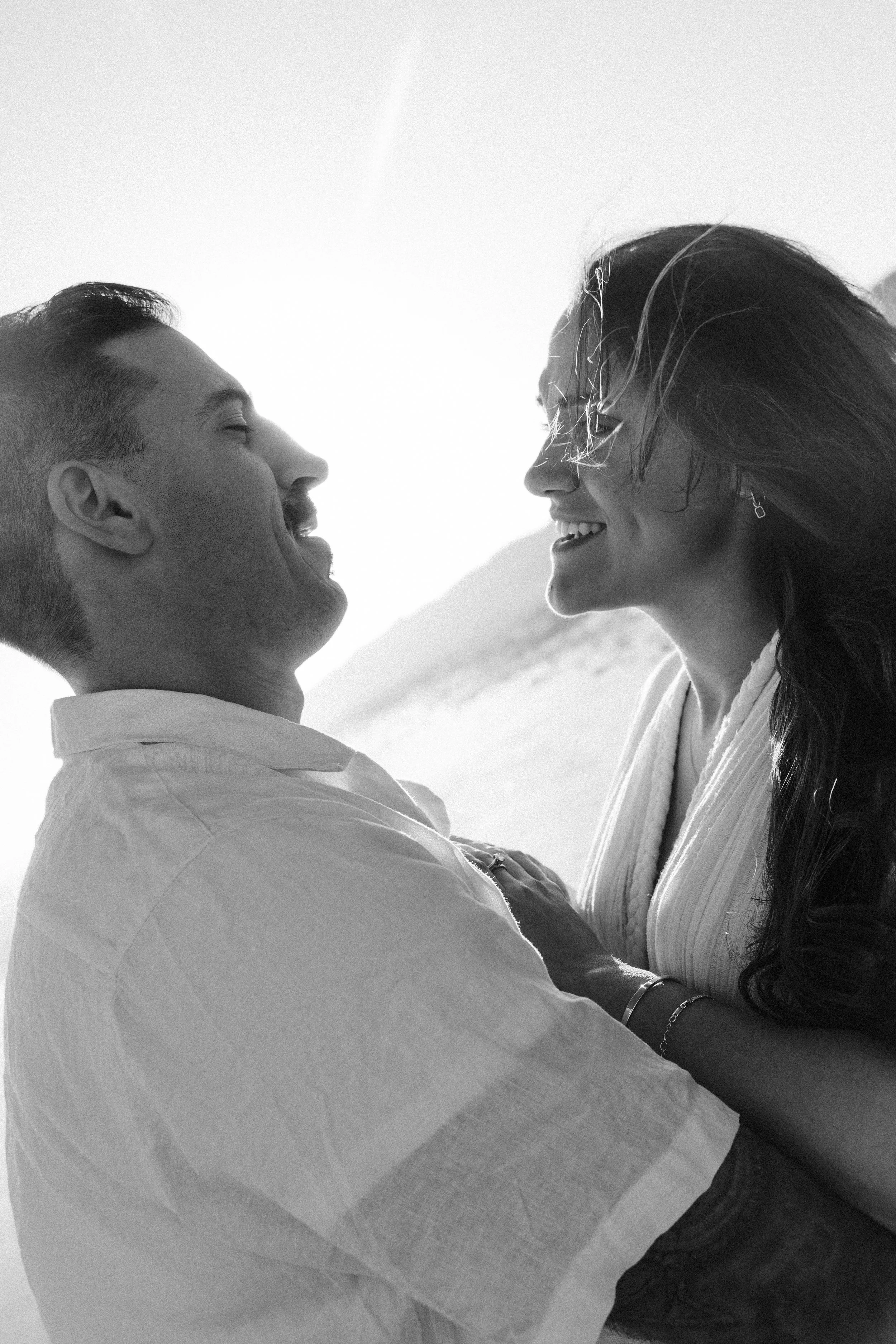 A black and white photo of a smiling couple outdoors, facing each other, enjoying a moment together with a background of what appears to be a beach or open landscape.