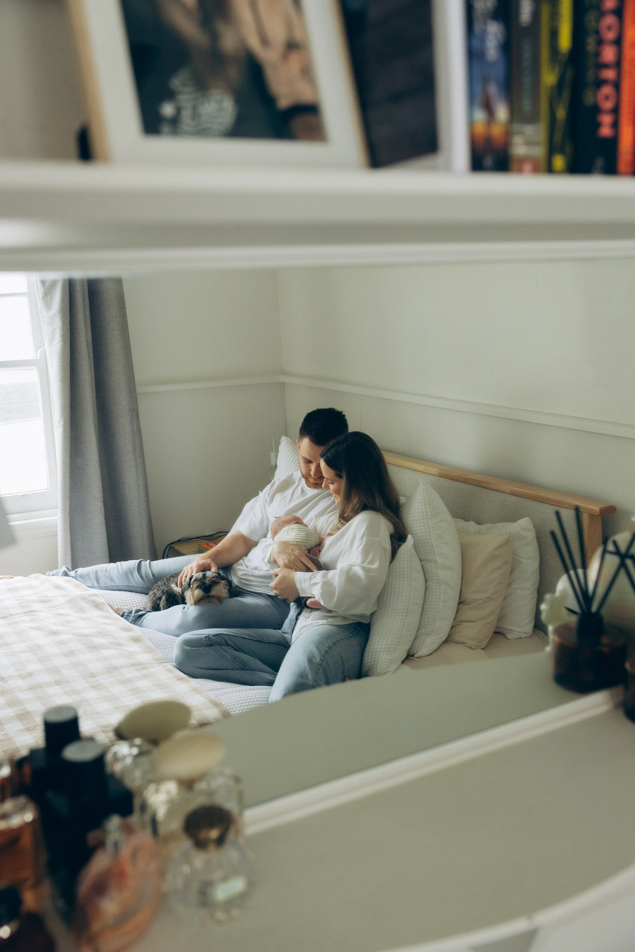 A couple sitting on a bed, breastfeeding a newborn, with a cat resting nearby, in a softly lit bedroom.