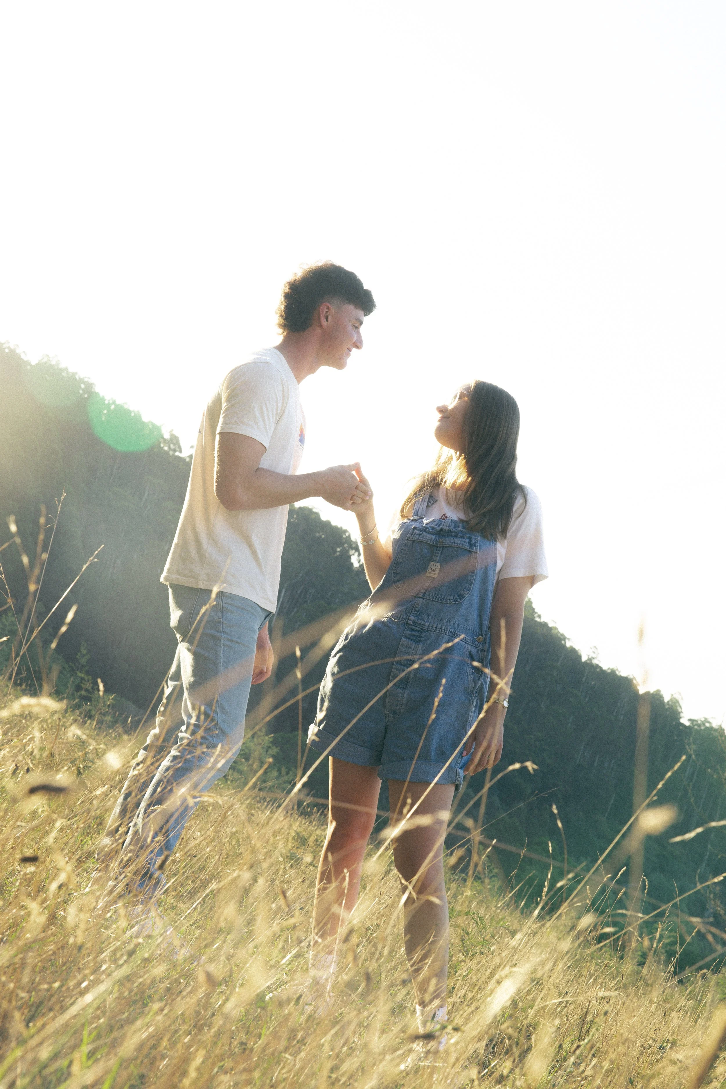 A young couple holding hands in a grassy field with a mountain in the background during sunset.