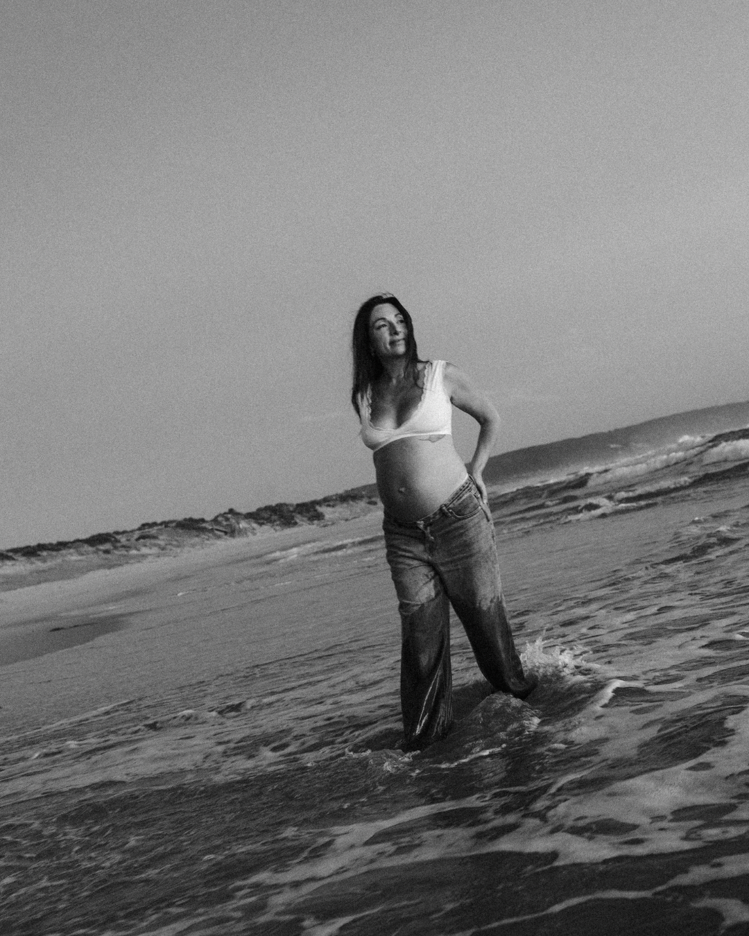 A woman standing in the ocean waves at the beach, wearing a white top and ripped jeans.