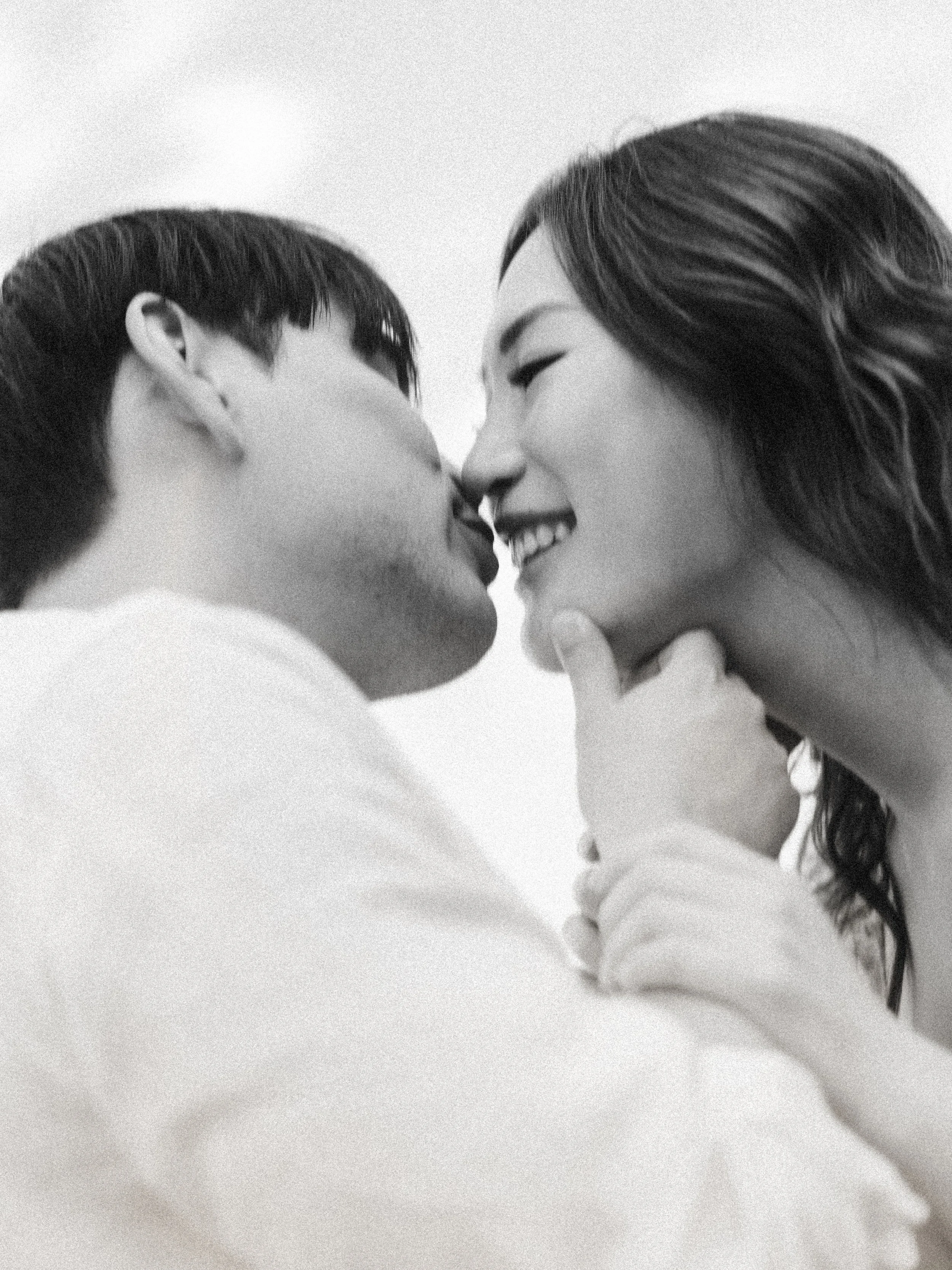 A black and white photo of a couple close together, about to kiss, with the woman smiling and the man's face partially visible.