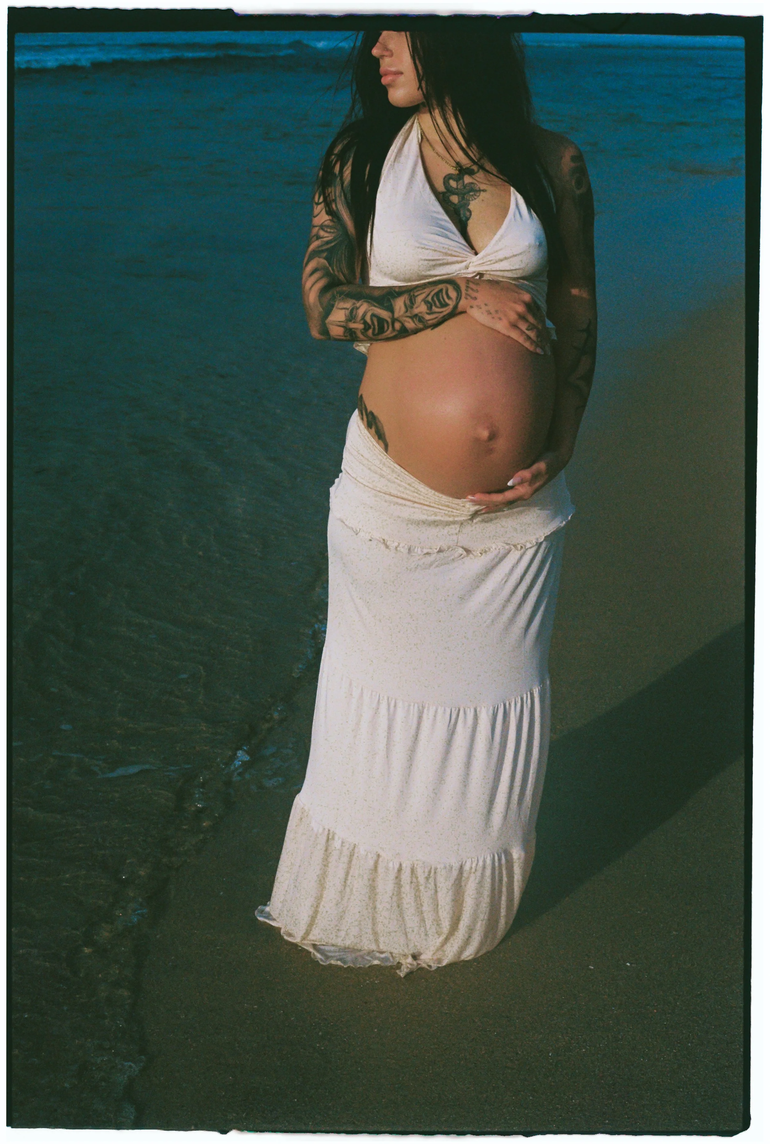 Pregnant woman with tattoos standing on the beach near the water, holding her belly.