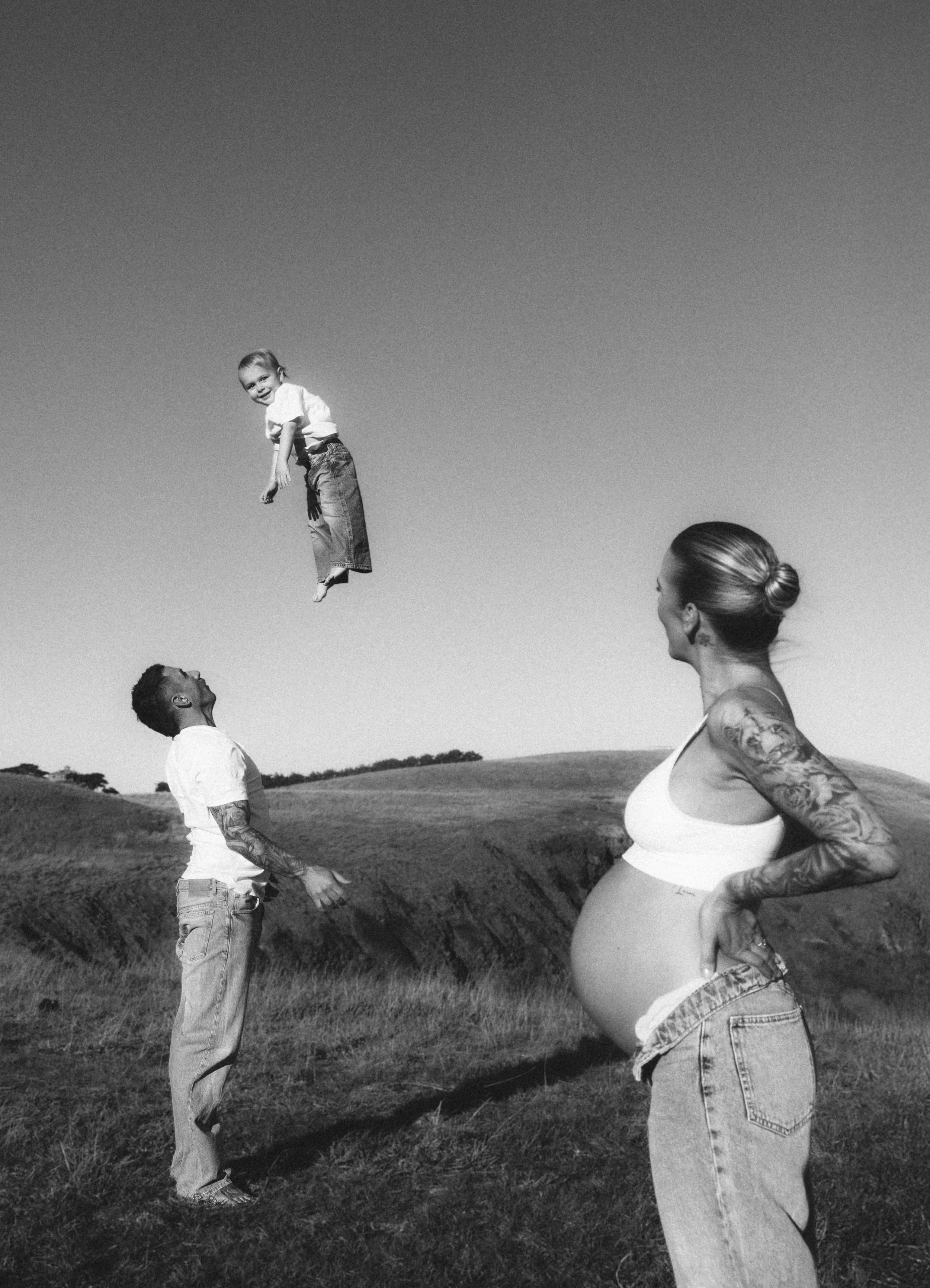 A family outdoors with rolling hills in the background. A pregnant woman with tattoos on her arms stands in the foreground, smiling and looking at a man and a girl. The man, also tattooed, is tossing the girl into the air, and she is smiling.