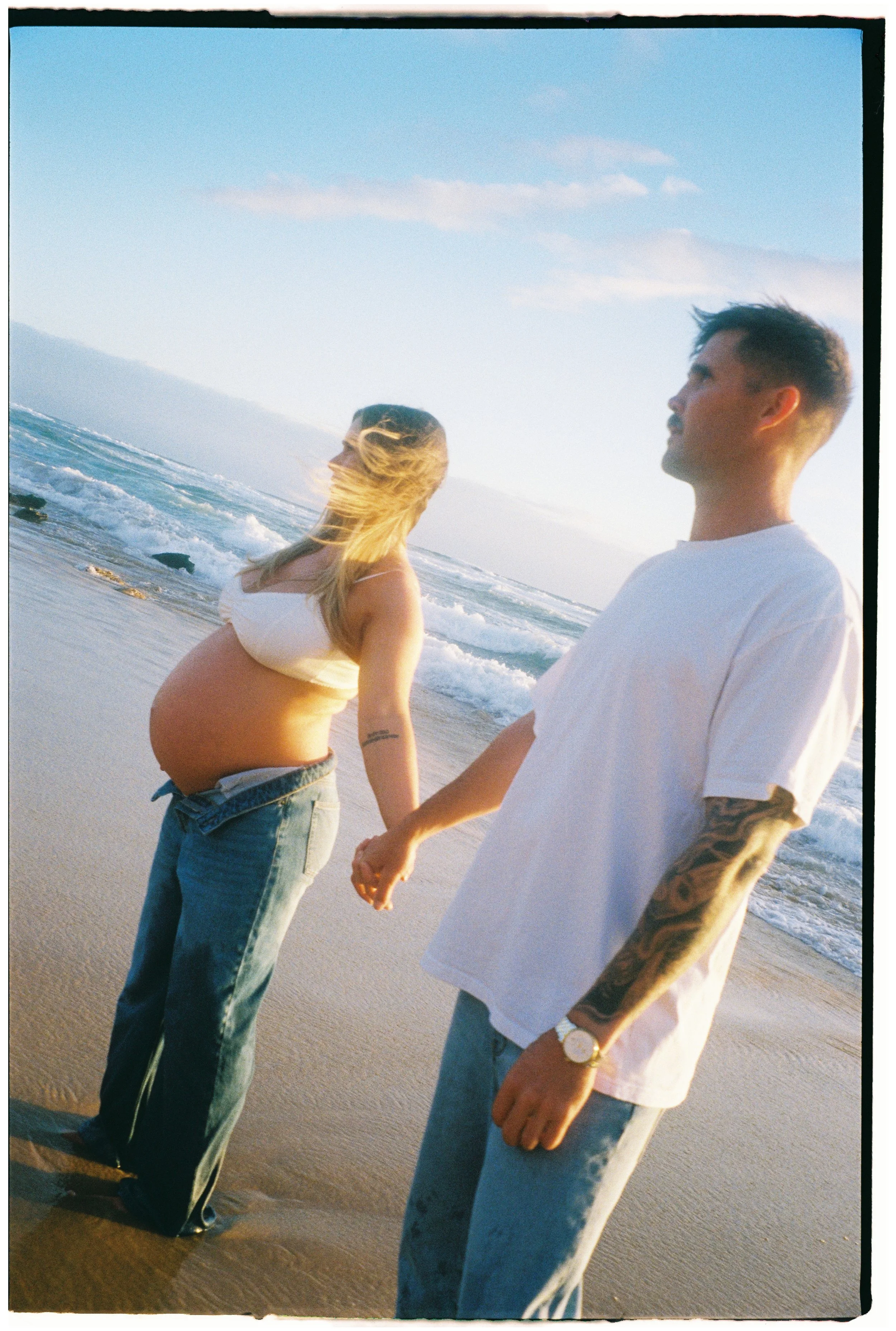 A pregnant woman and a man holding hands on a beach during sunset, with ocean waves in the background.