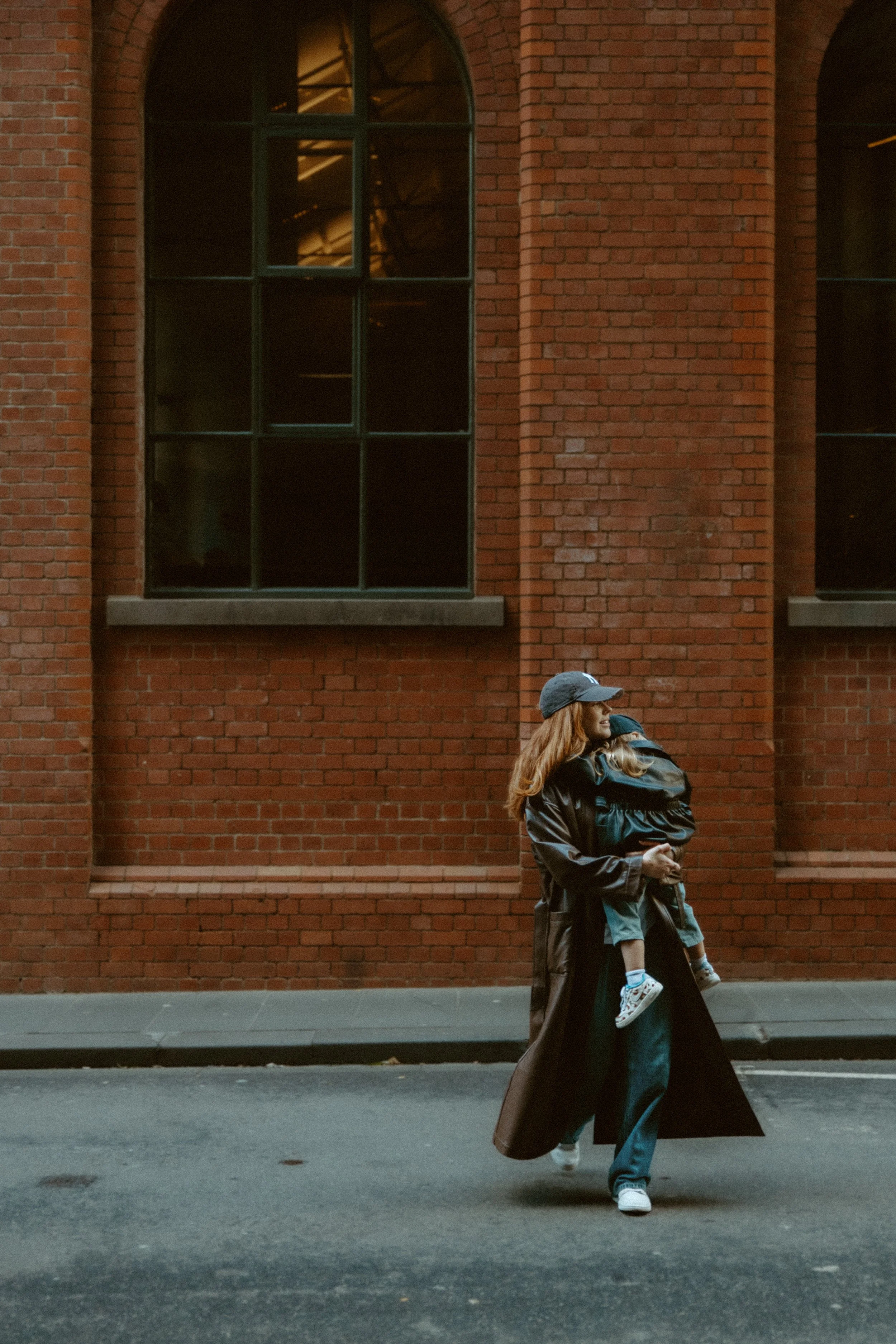 A woman wearing a dark coat and a baseball cap walks on a city street, carrying a child in her arms. The background features a brick building with large arched windows.