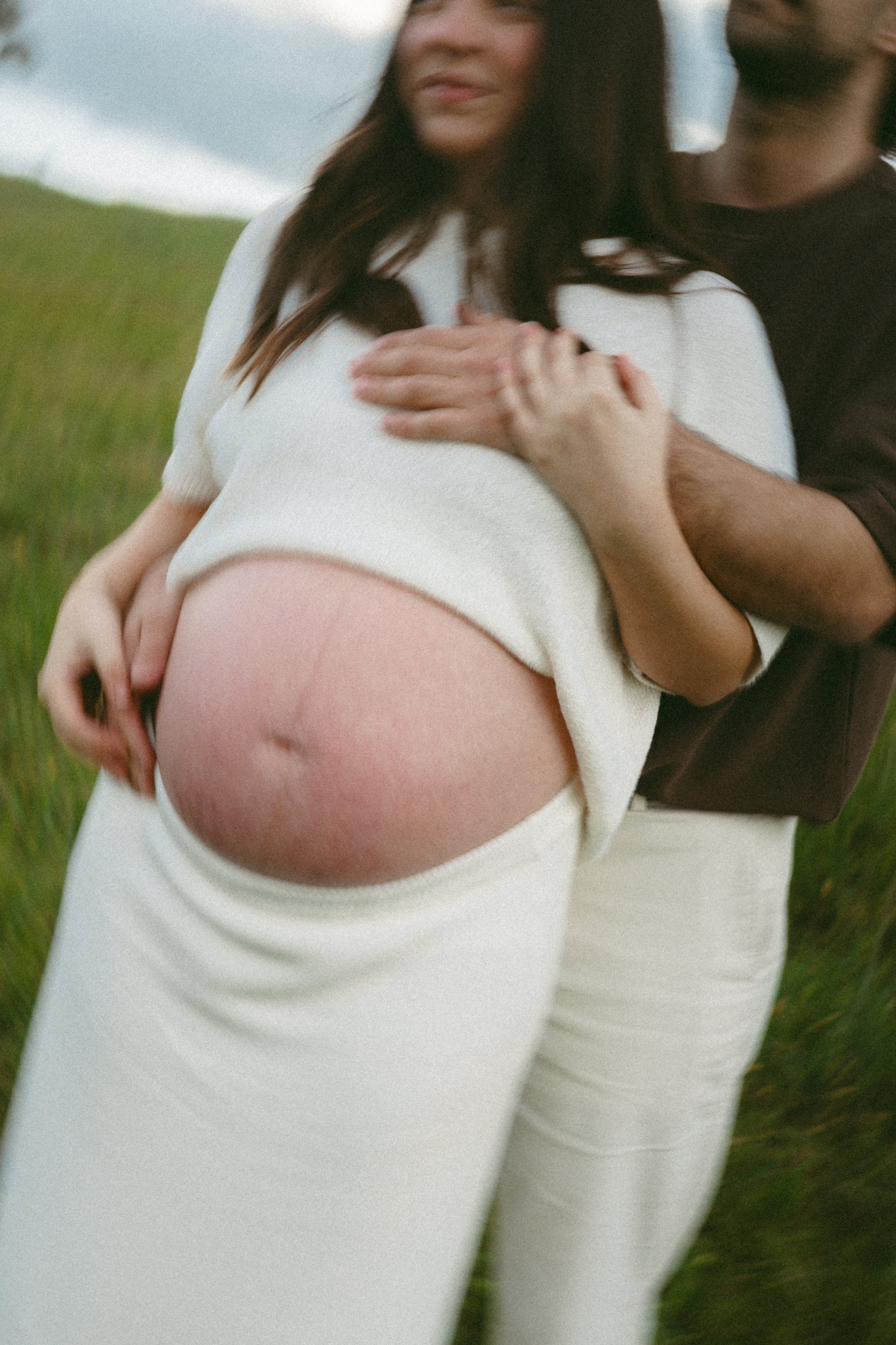 A pregnant woman wearing a white outfit holding her belly outdoors with another person supporting her, in a grassy field.