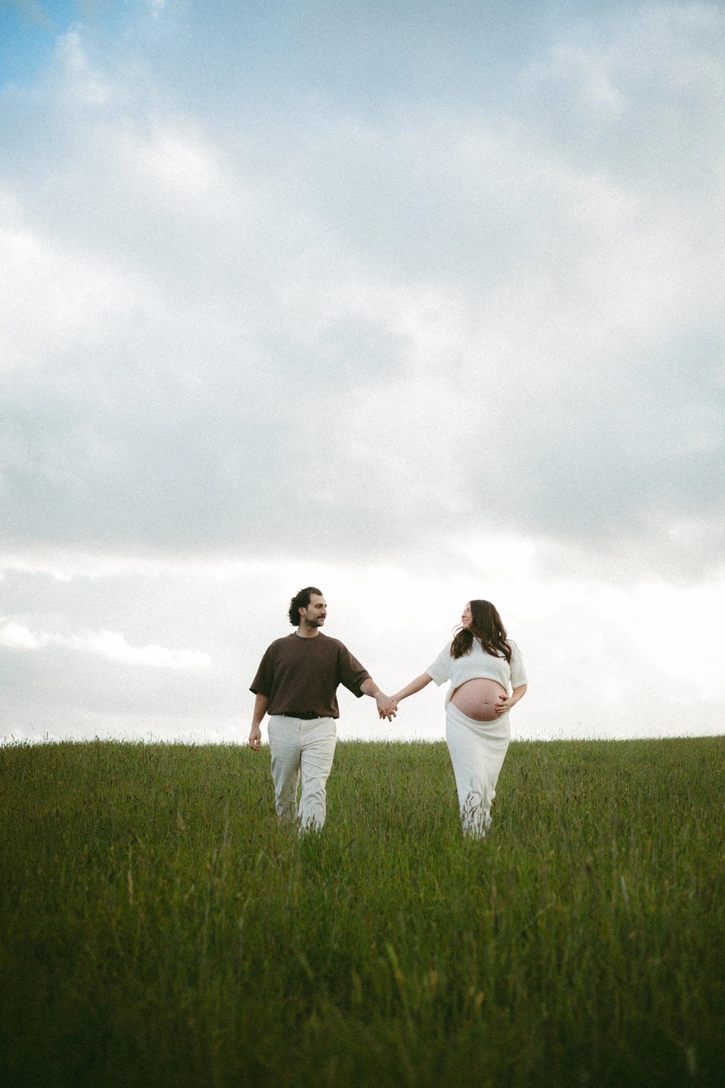 A pregnant woman and her partner holding hands and walking in a grassy field under a cloudy sky.