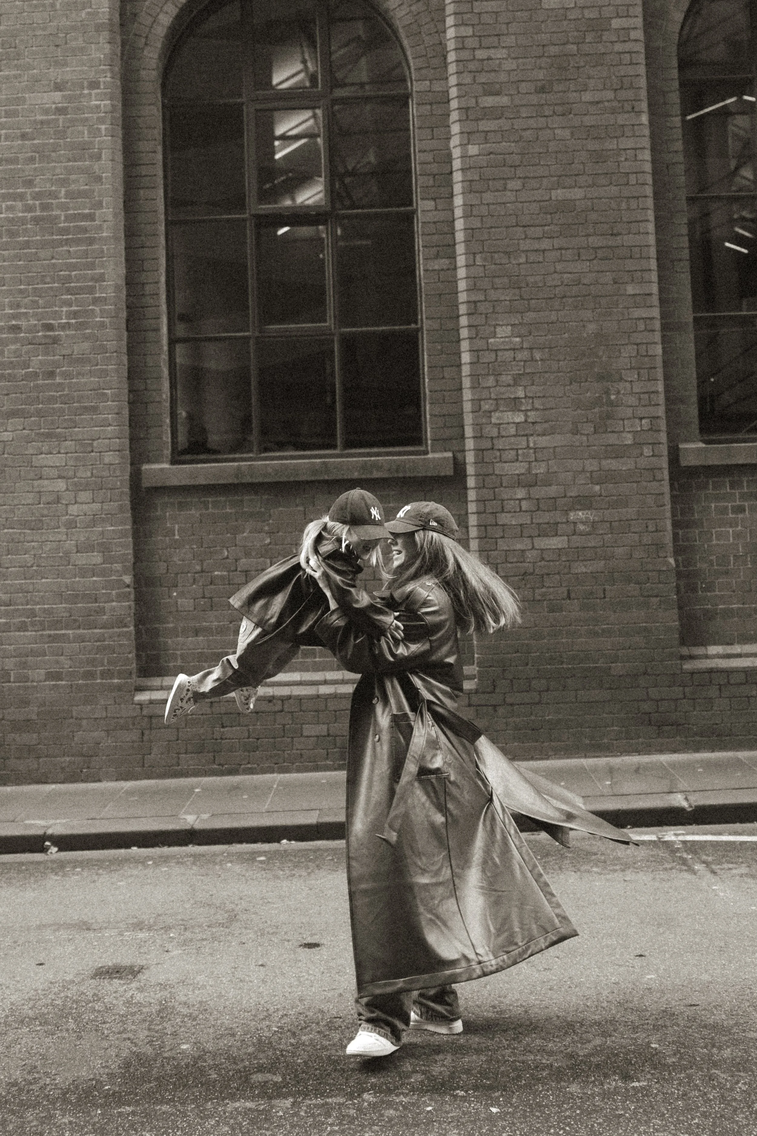 Two women, one lifting the other, are smiling and wearing jackets and baseball caps, standing outside on a city street at night with a brick building and large windows in the background.