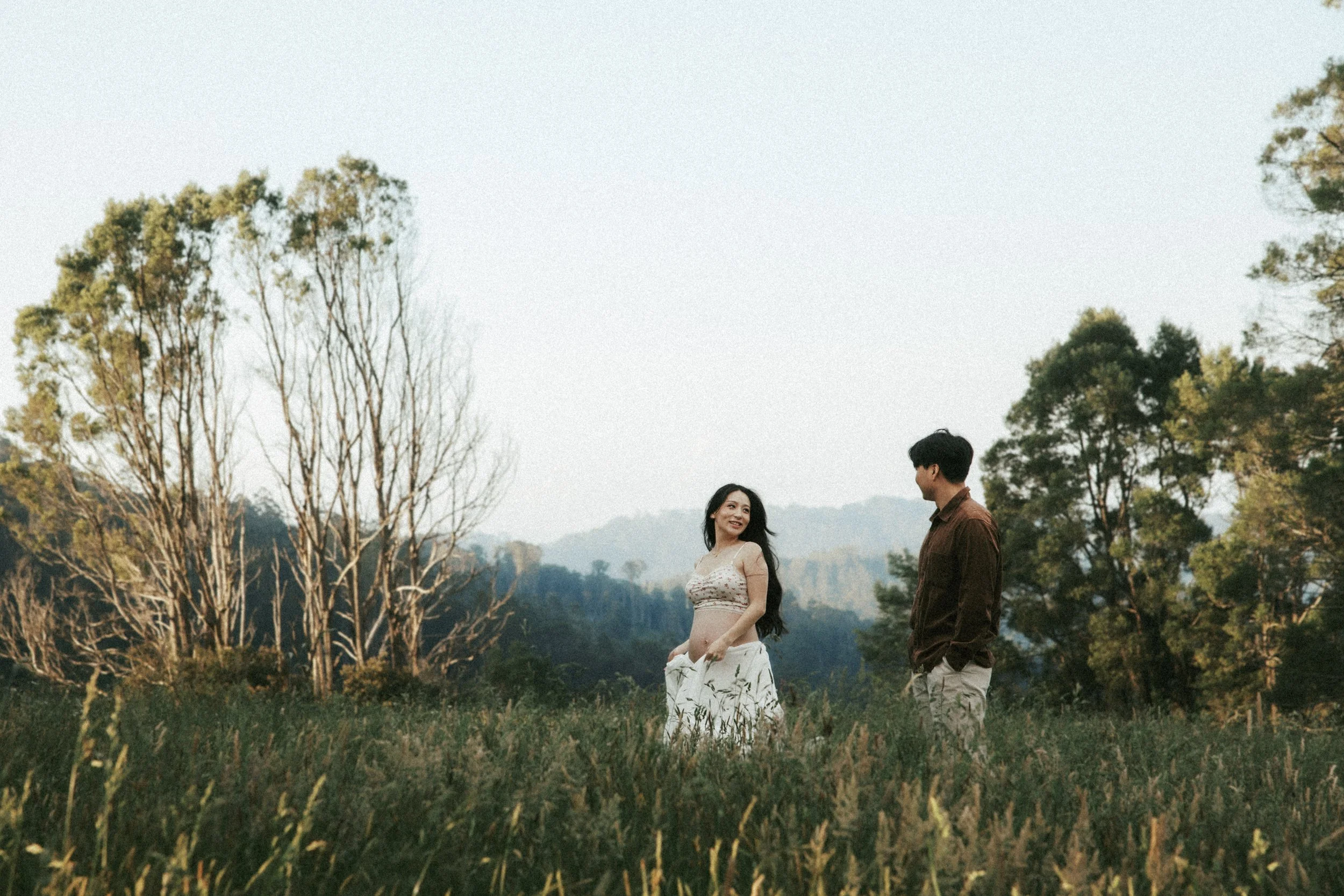 A couple standing in a grassy field with trees and mountains in the background, the woman wearing a sleeveless top and skirt, and the man wearing a brown shirt and light-colored pants, smiling and looking at each other.