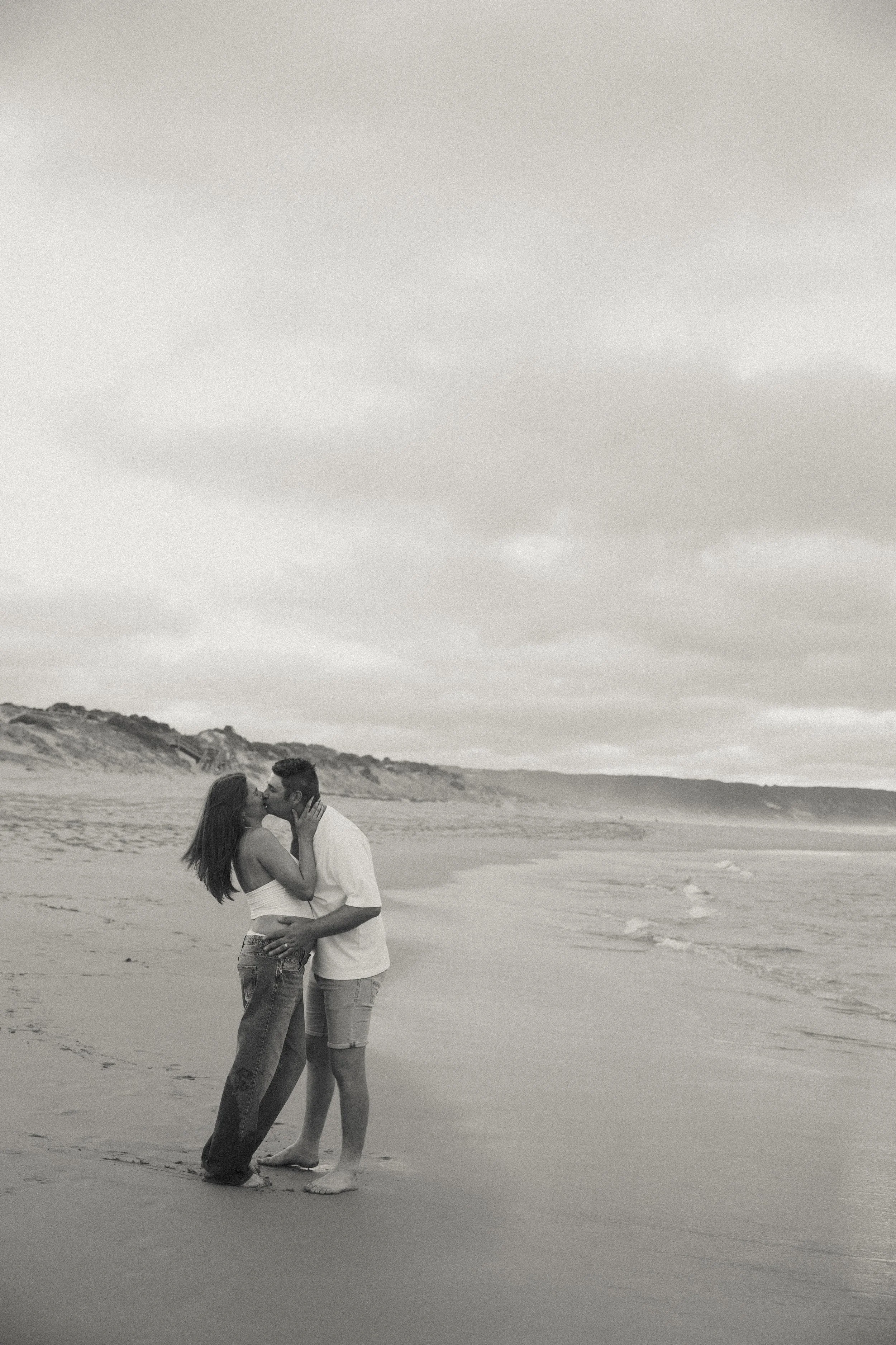 A couple kissing on the beach, holding each other close, with ocean waves and cloudy sky in the background.