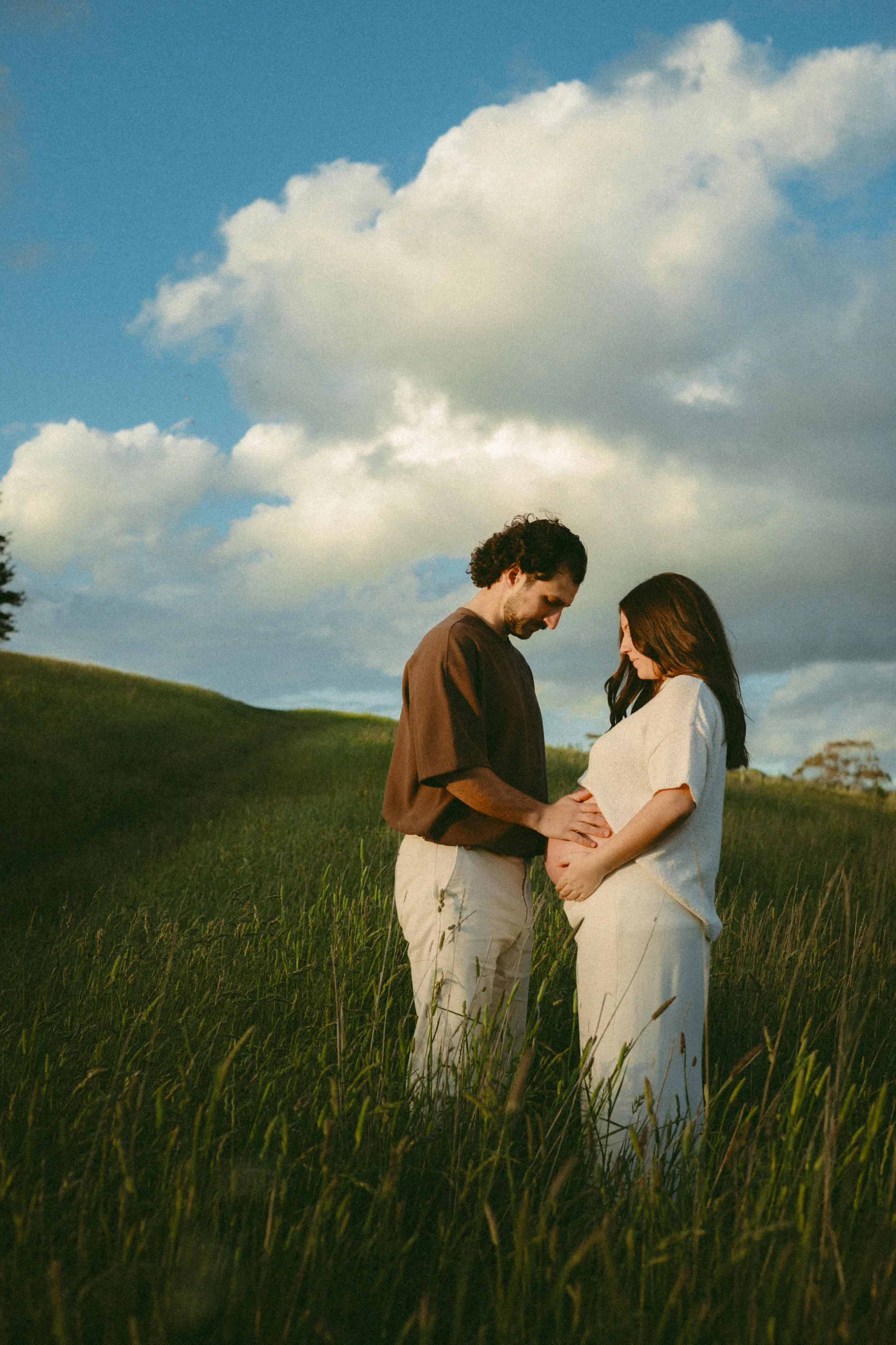 A pregnant woman and her partner standing in a grassy field under a blue sky with clouds, touching her baby bump.