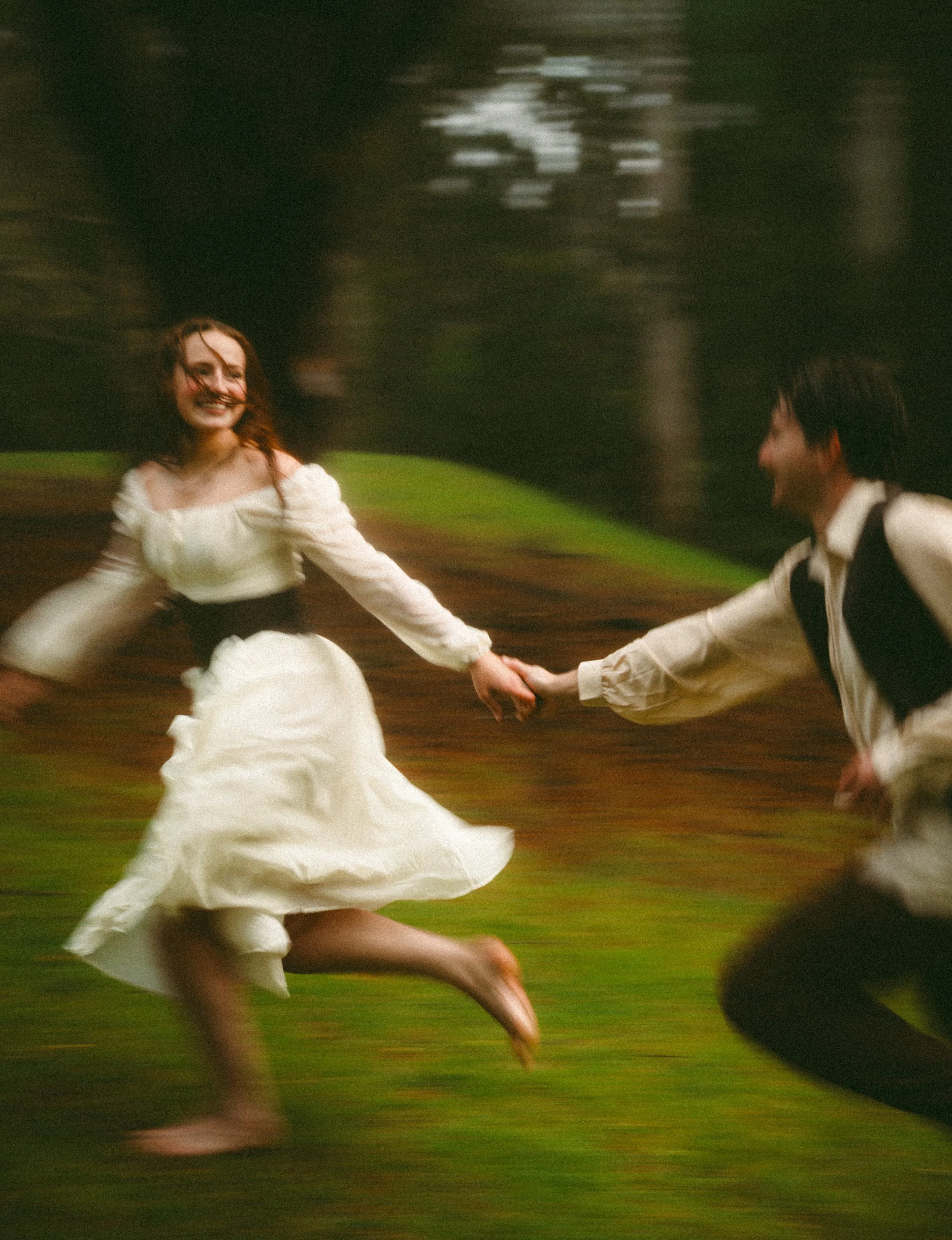 A woman in a white dress and a man in a white shirt and black vest are holding hands and spinning in a grassy outdoor area with trees, creating a motion blur.