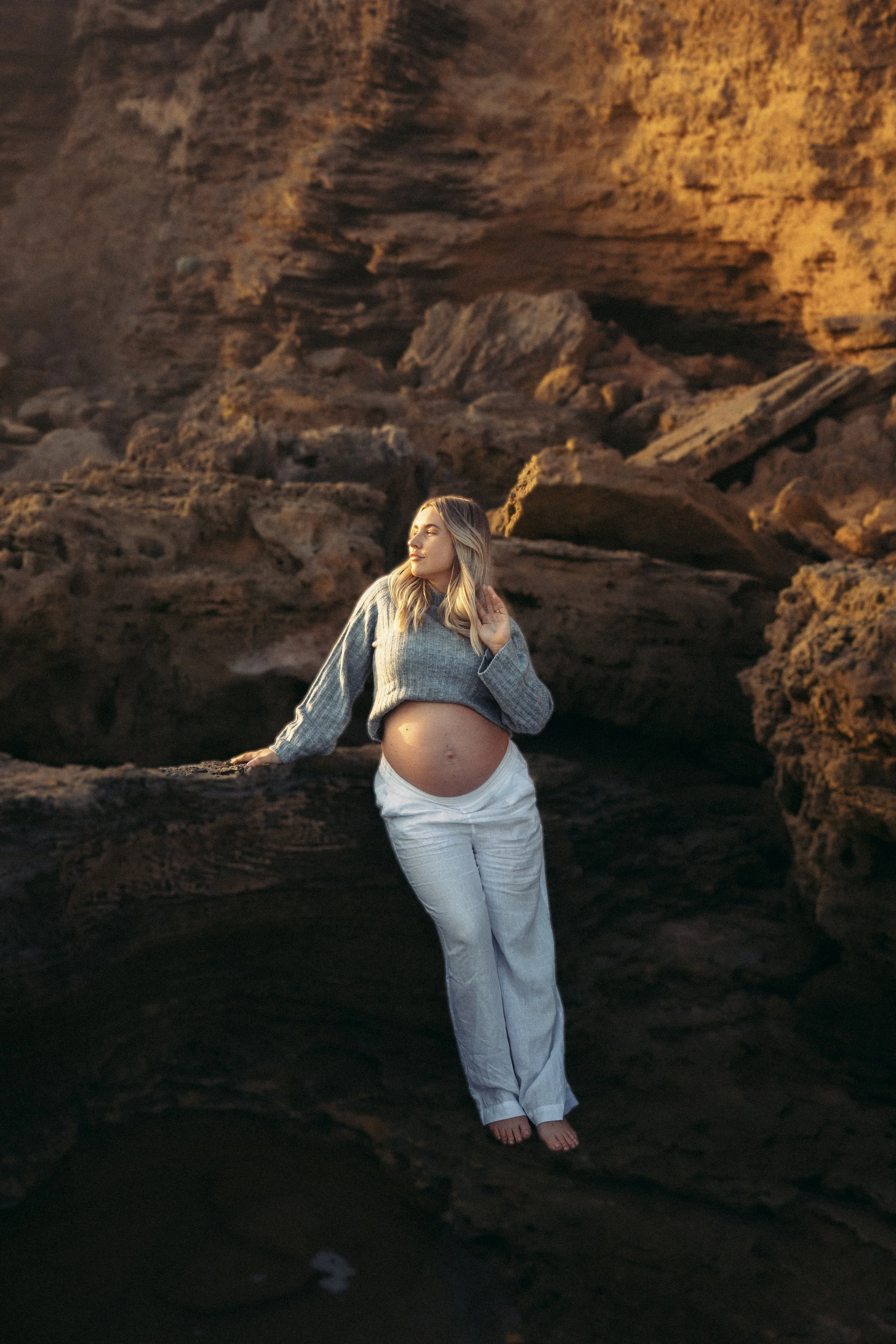 A pregnant woman standing on rocks at a beach, wearing a gray sweater and white pants, with her hand on her belly and looking to the side.