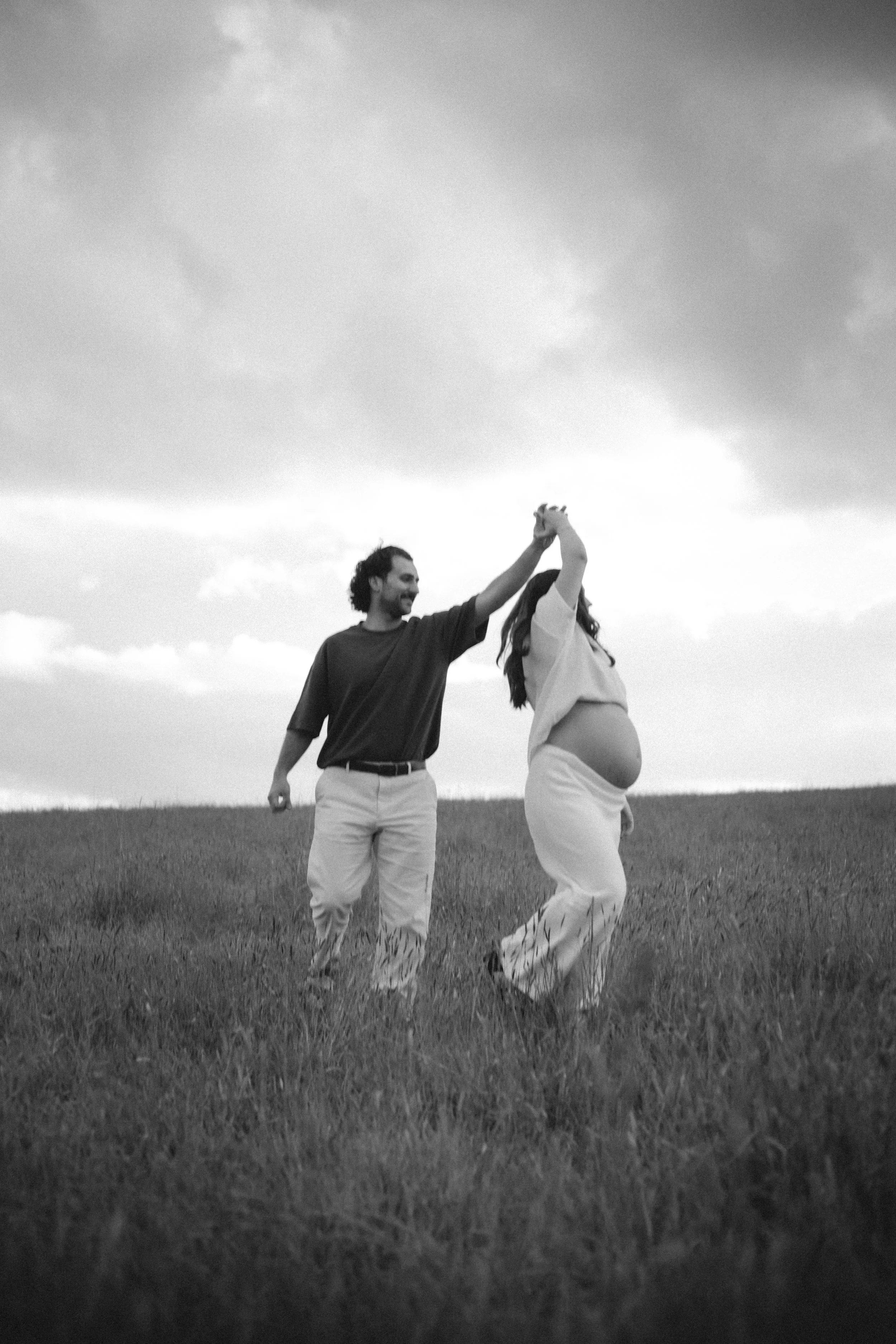 A man and a pregnant woman holding hands and dancing in a grassy field, with a cloudy sky in the background, in black and white.