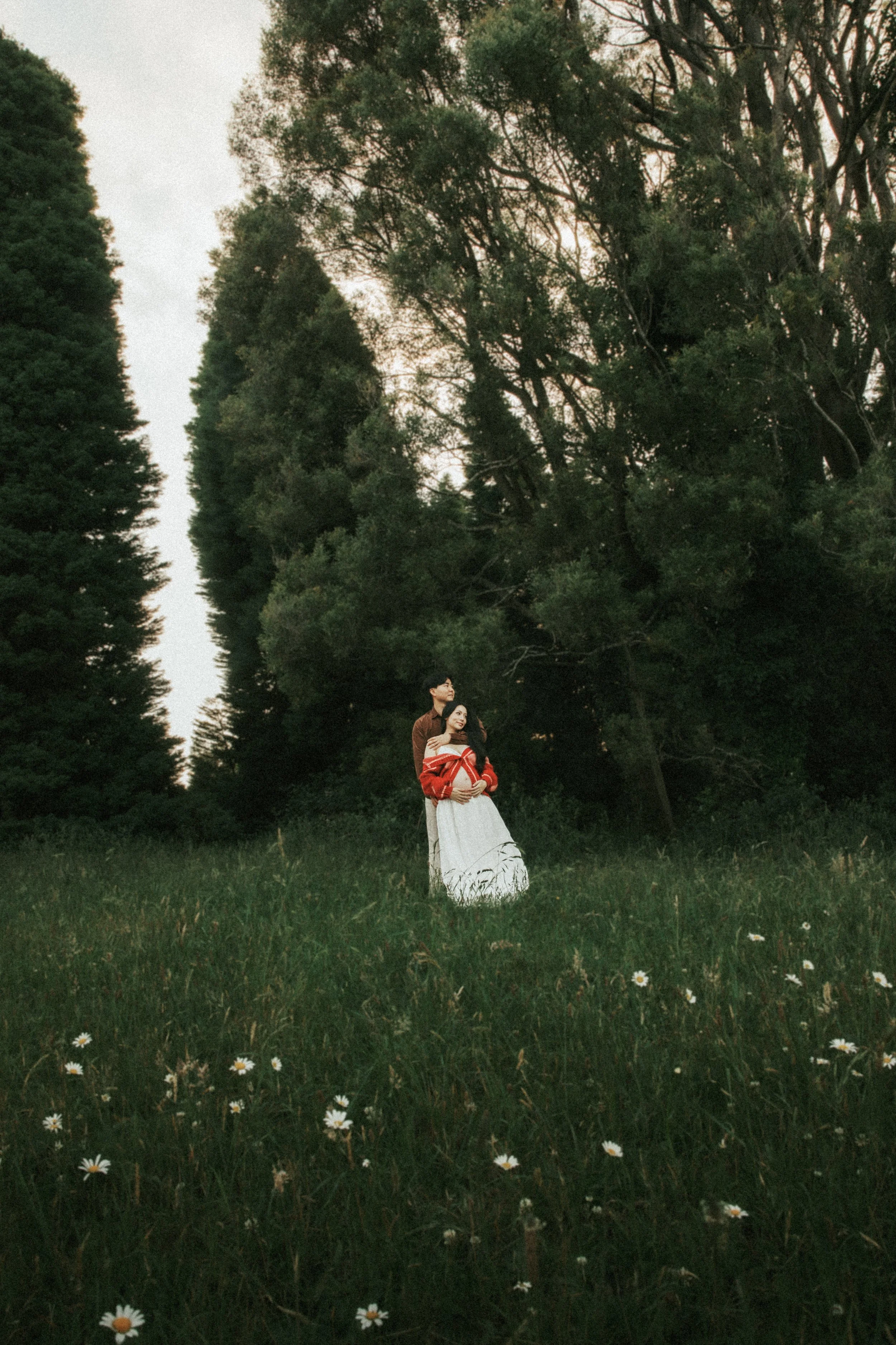 Couple standing in a grassy field with tall trees in the background, during sunset. The woman is wearing a white skirt and a red off-the-shoulder sweater, and the man is wearing a brown jacket.