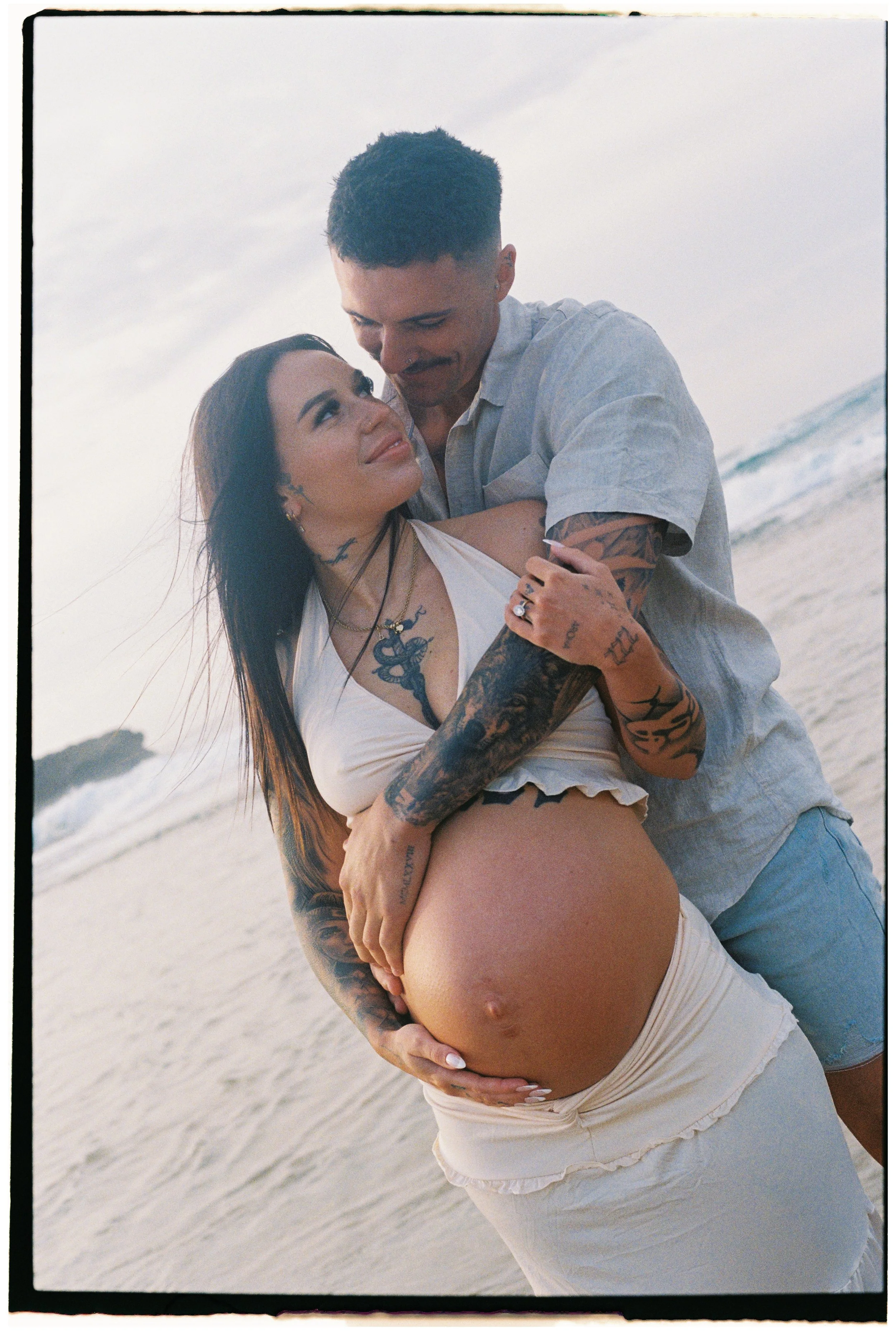 A pregnant woman and a man on the beach, embracing each other. The woman has tattoos and is wearing a white top and skirt, and the man has tattoos and is wearing a light-colored shirt and shorts. They are smiling and looking at each other, with the ocean in the background.
