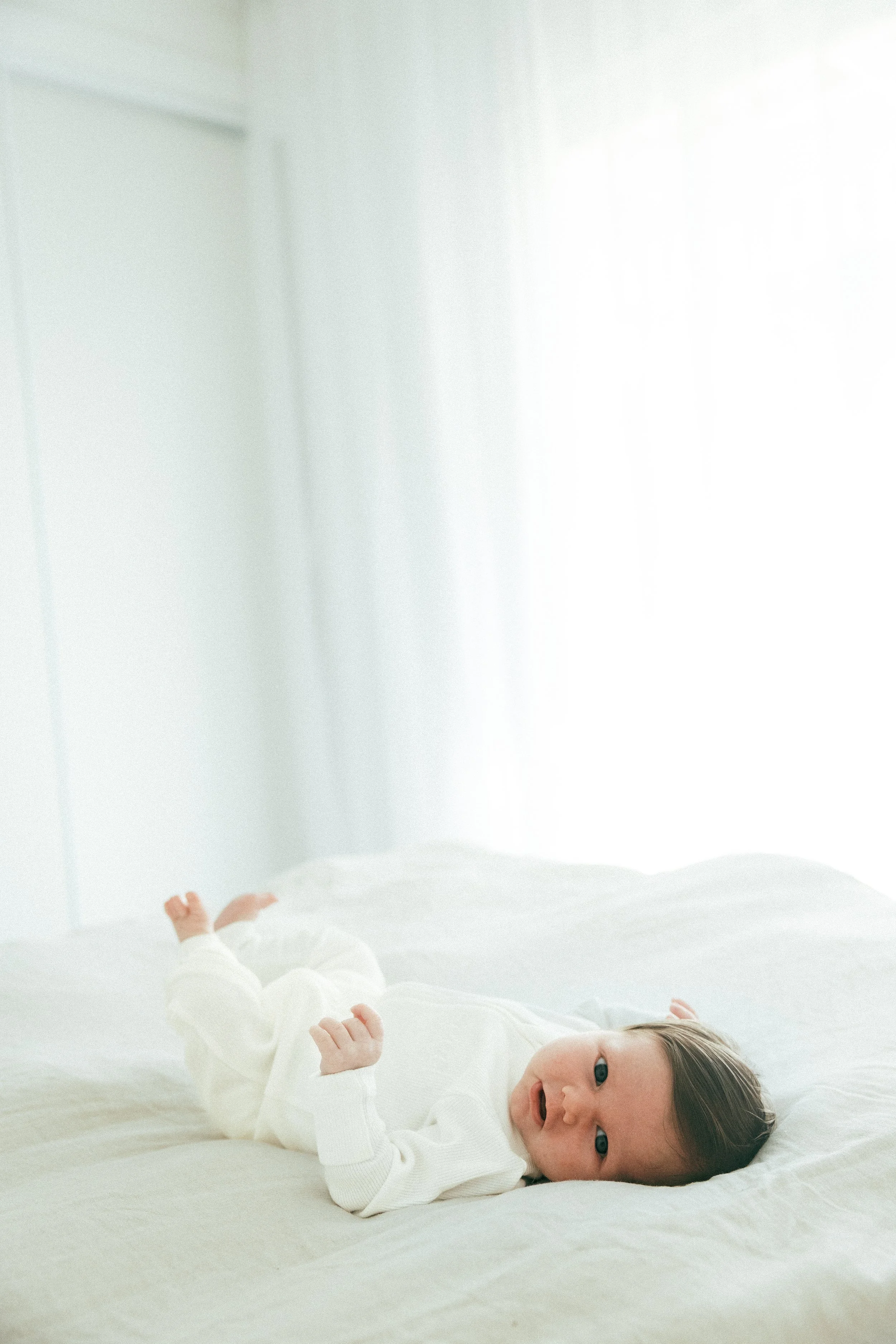 A baby laying on a white bed in a bright, softly lit room with white curtains.