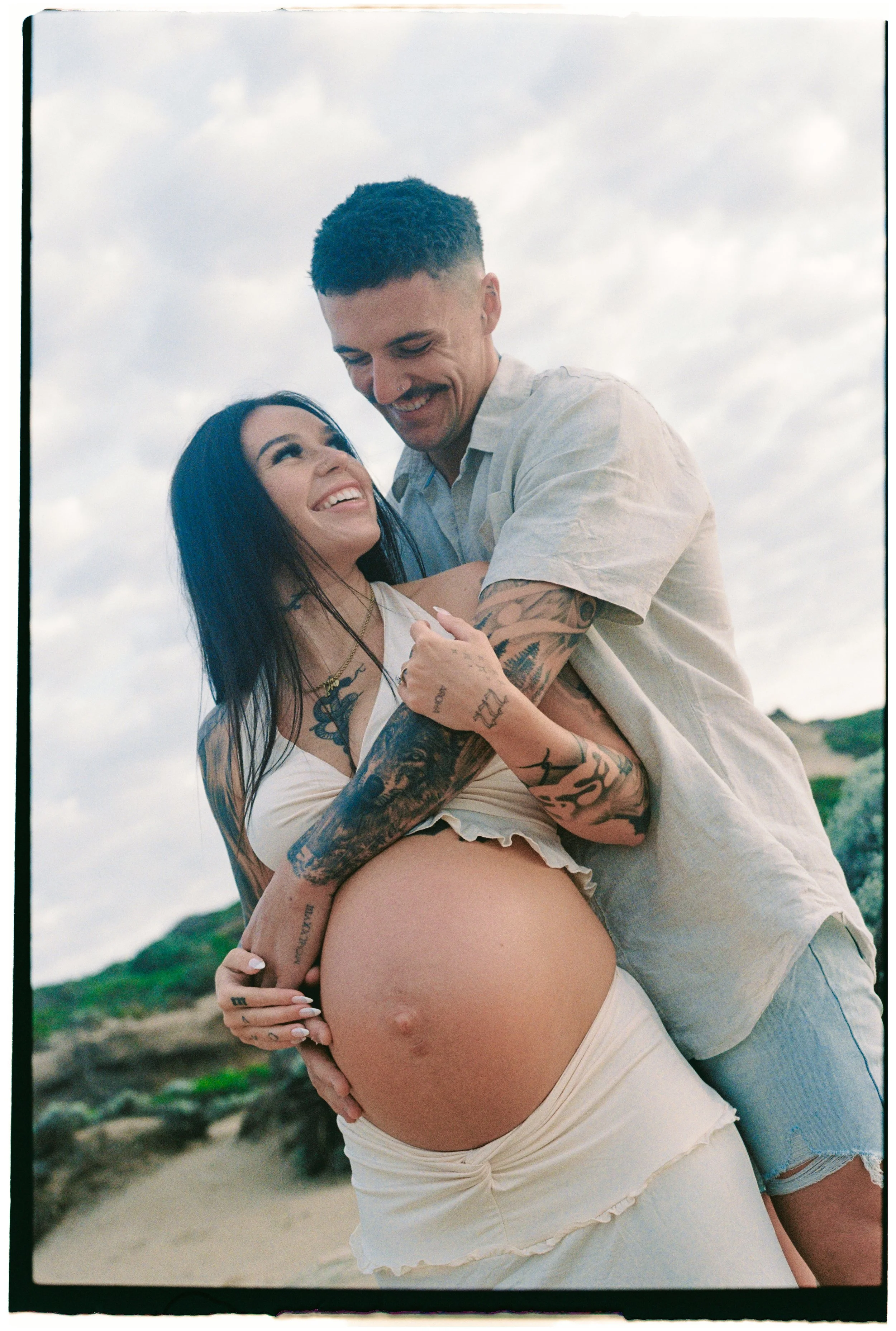 A smiling pregnant woman with tattoos on her arms and chest, standing outdoors on a beach, being embraced by a man who is also smiling. The man is wearing a light-colored shirt and shorts.