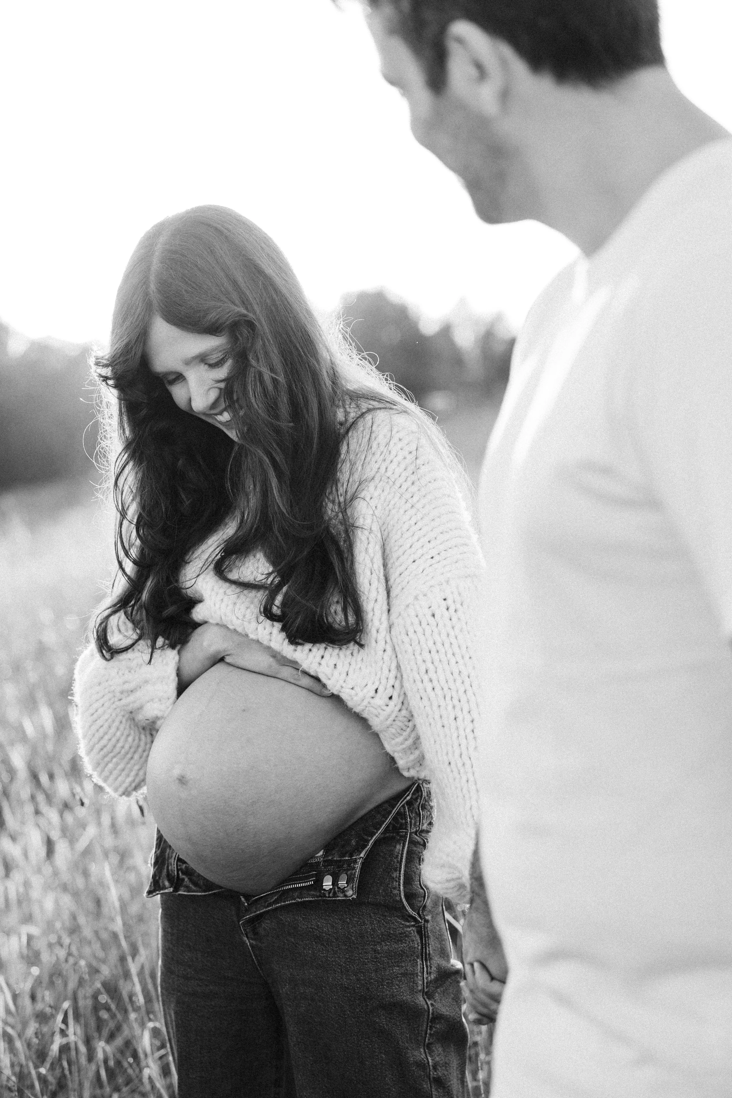 Black-and-white photo of a pregnant woman smiling and looking down, holding her belly, with a man standing nearby in an outdoor field.