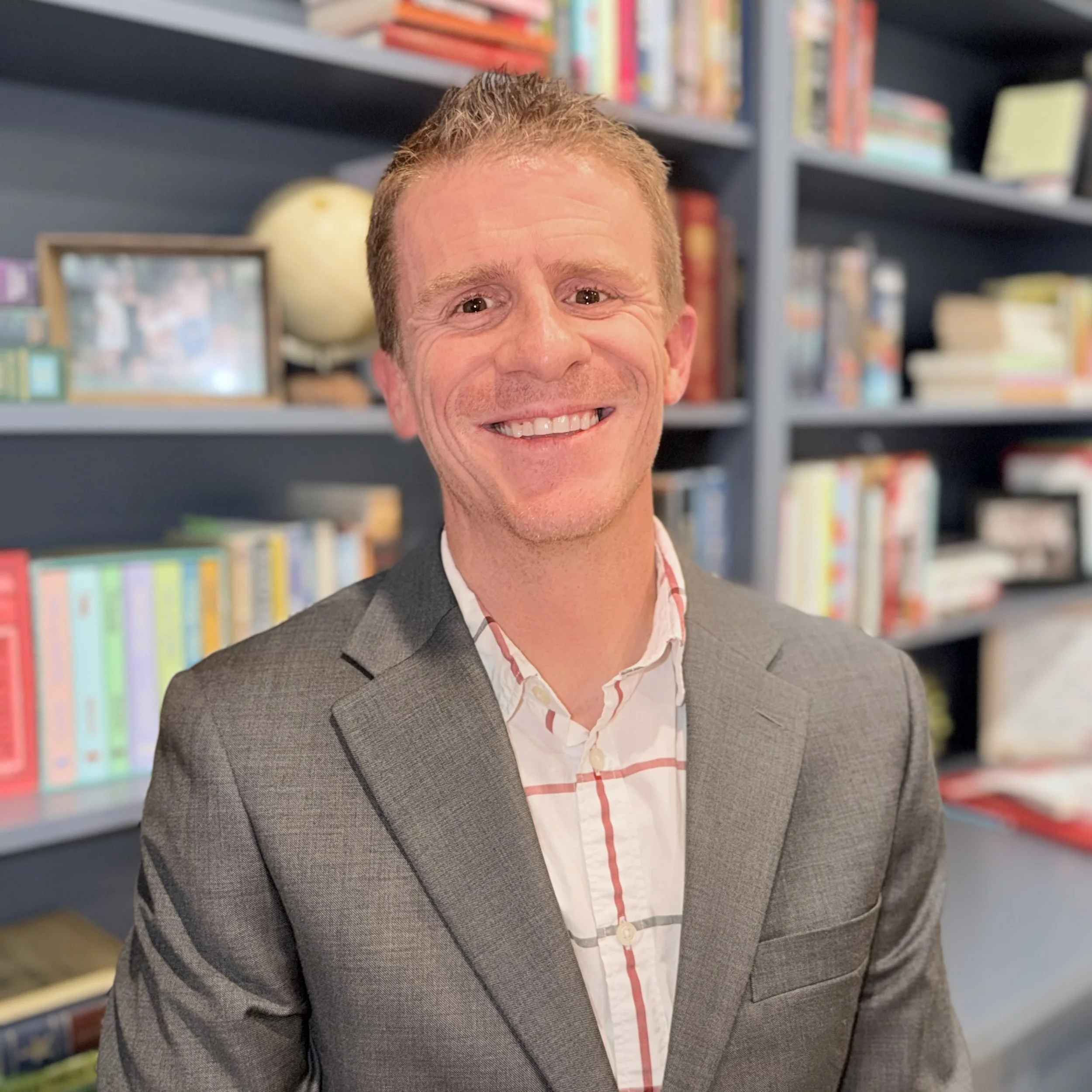 A smiling man in a gray suit with a white shirt featuring a red and black grid pattern, standing in front of a bookshelf filled with books and a globe.