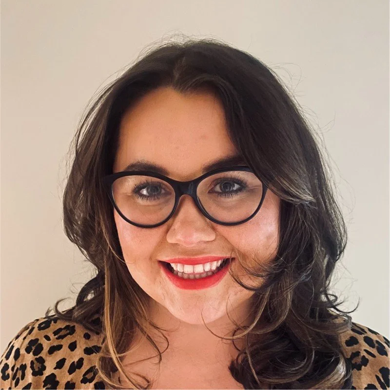 A woman with dark, wavy hair, wearing glasses and leopard print top, smiling against a plain background.