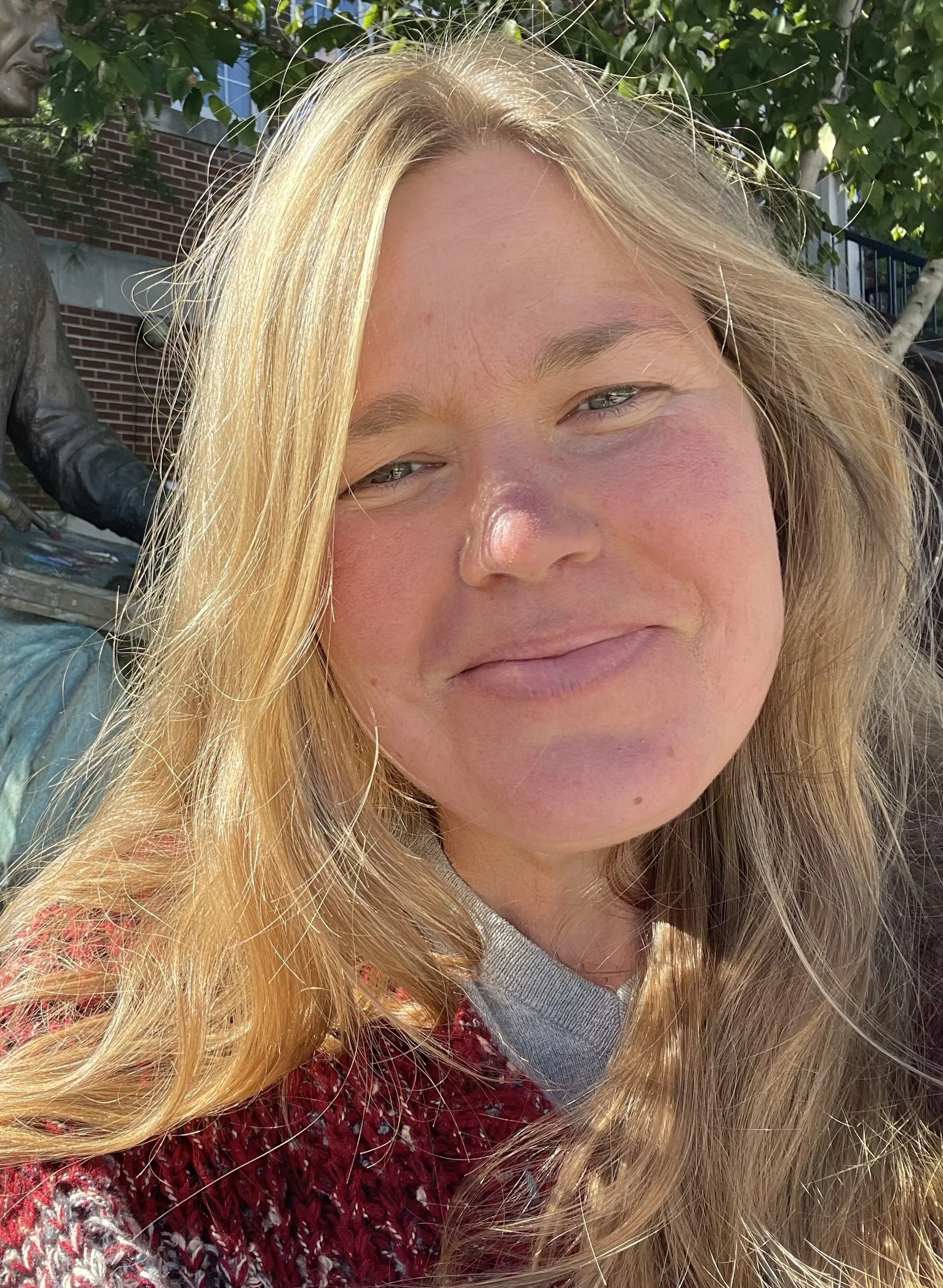 Close-up of a woman with long blonde hair, smiling, outdoors with trees and a brick building in the background.