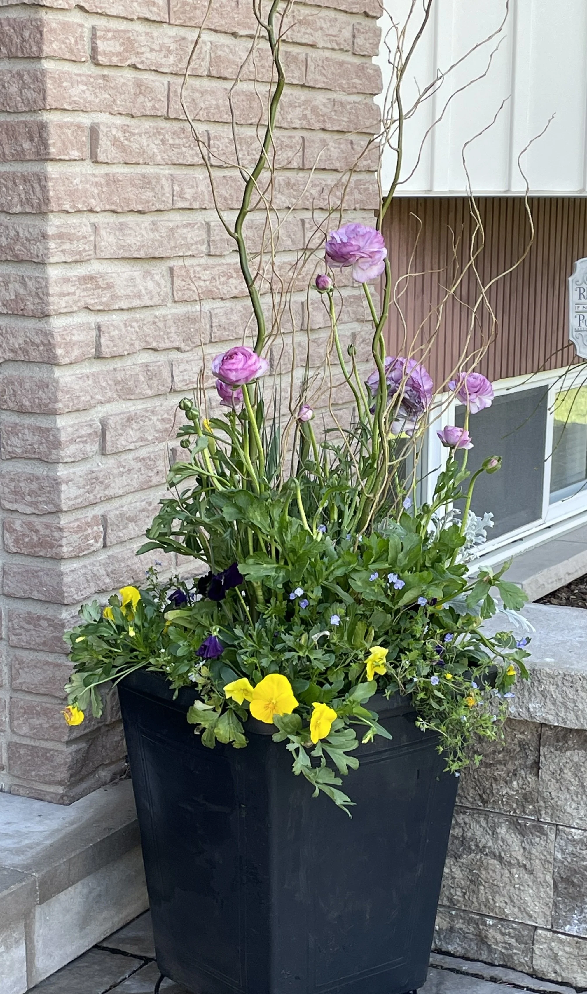 A black planter with various colorful flowers, including pink, purple, and yellow, placed on a stone patio against a brick wall and window.