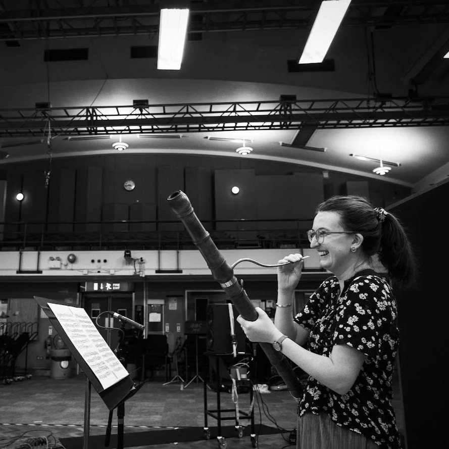 A woman with glasses and a ponytail smiling and holding a baroque bassoon in a rehearsal or performance space.